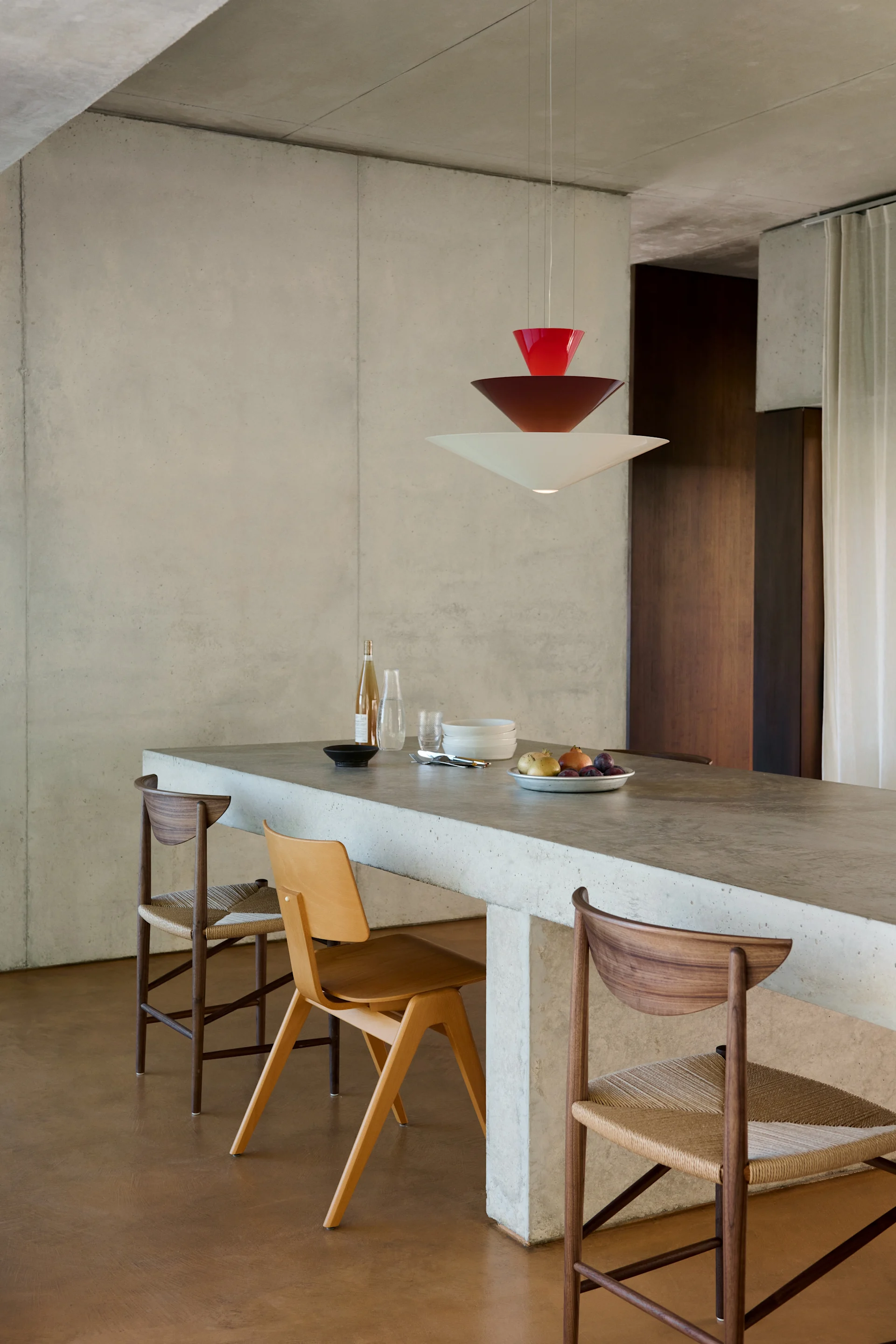 Salle à manger moderne avec îlot en béton, trois chaises en bois et un luminaire suspendu étagé rouge et blanc.