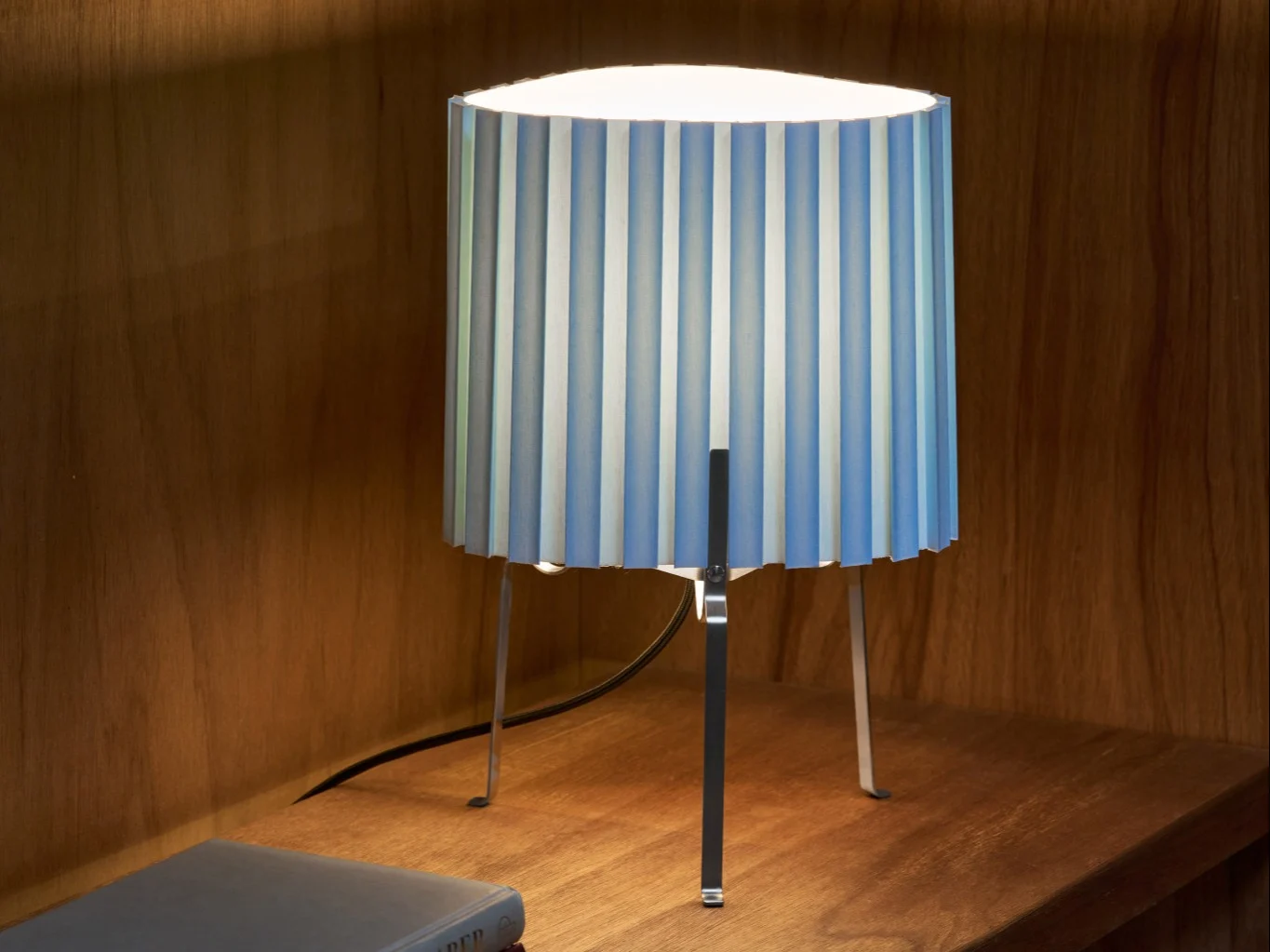 Lampe de table avec abat-jour plissé à rayures bleues et blanches sur une étagère en bois, éclairant la zone.