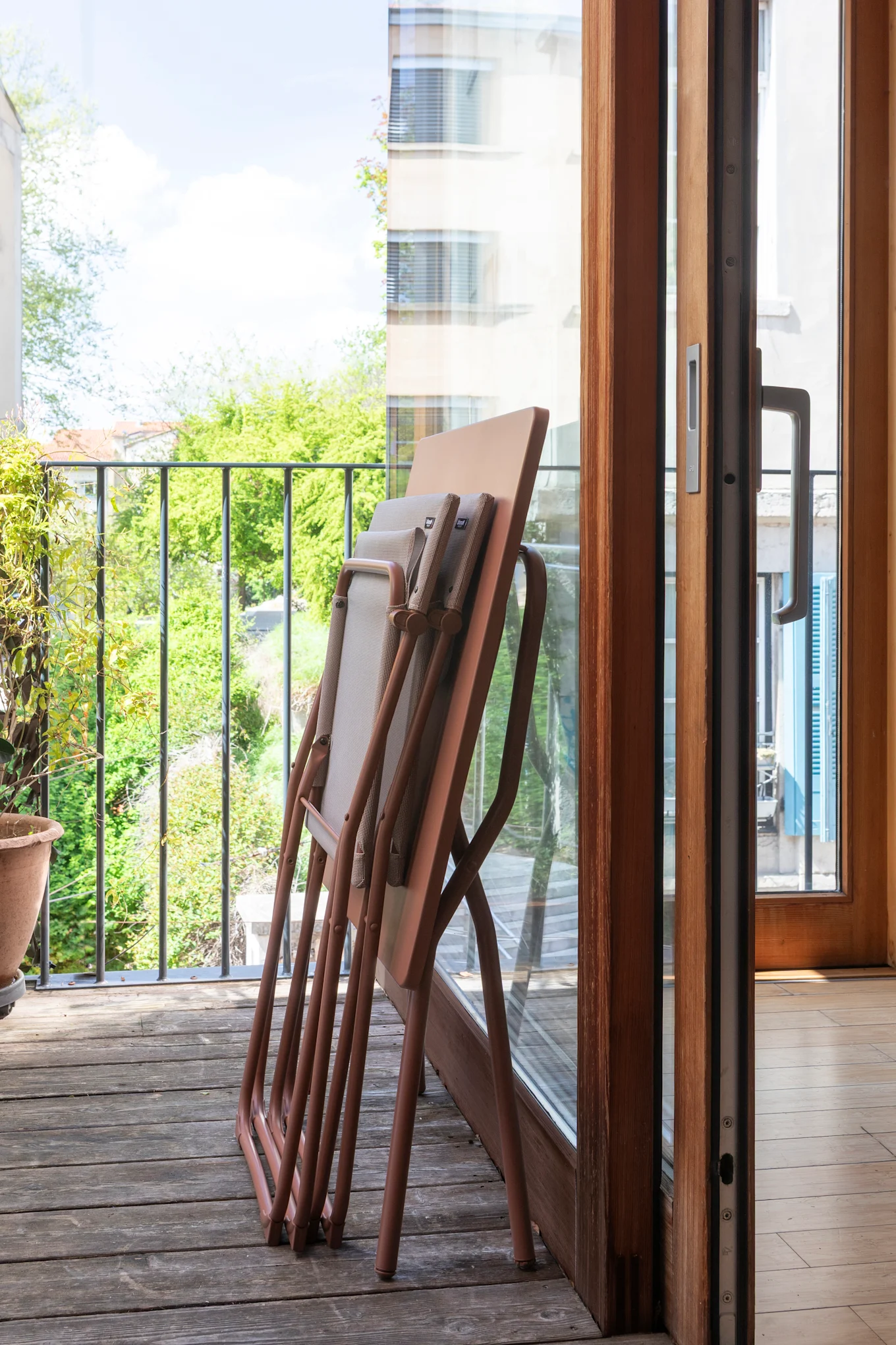 Chaises pliantes empilées sur le plancher en bois d'un balcon à côté d'une porte vitrée, donnant sur des arbres verts.