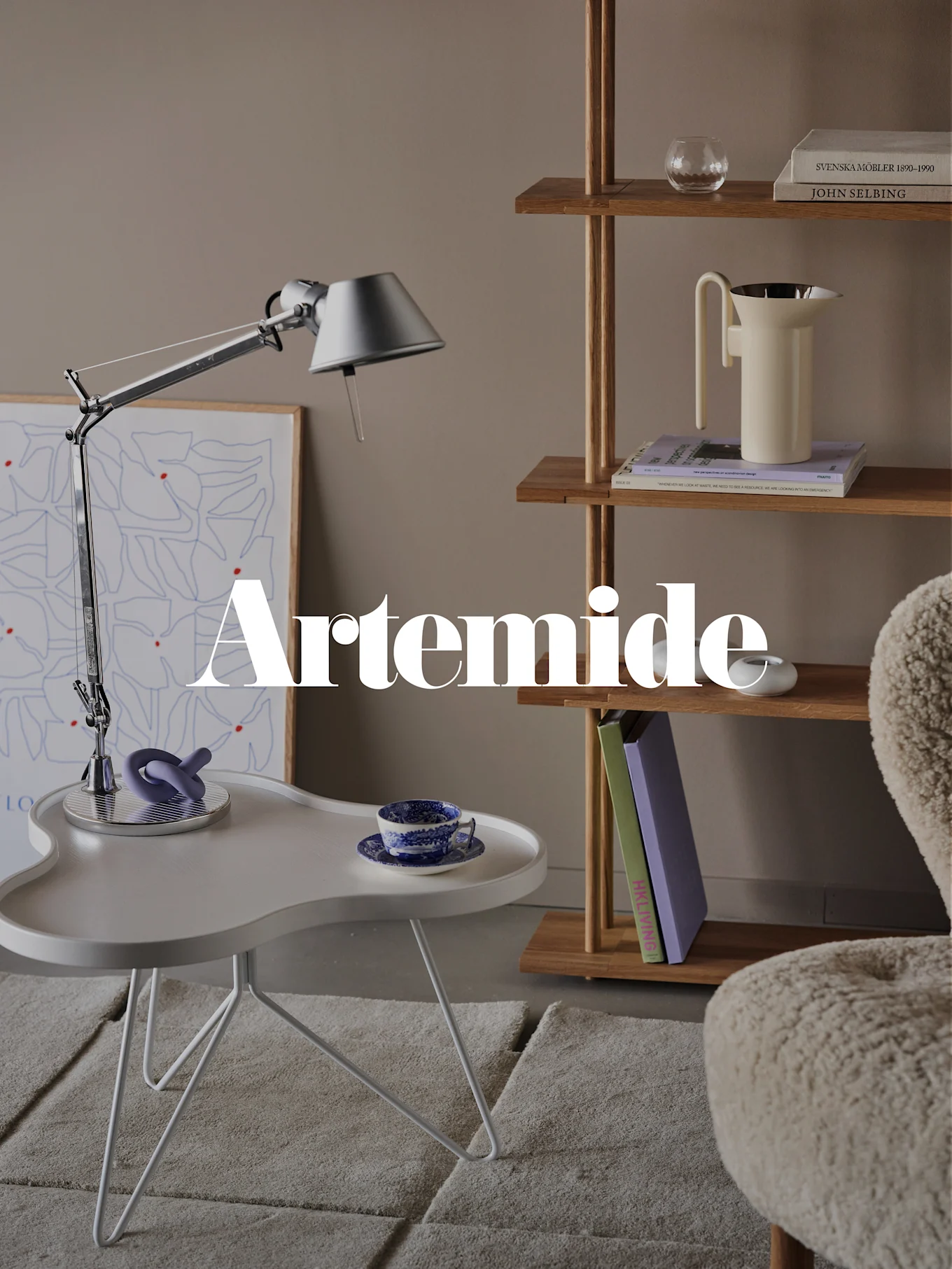 Un salon moderne avec une lampe de bureau argentée, une table d'appoint blanche, une bibliothèque en bois et un fauteuil crème. Superposition de texte : 'Artemide'.