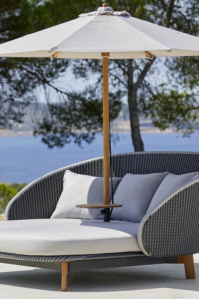 Chaise longue d'extérieur avec coussins gris clair et parasol assorti, au bord de la mer avec des arbres en arrière-plan.