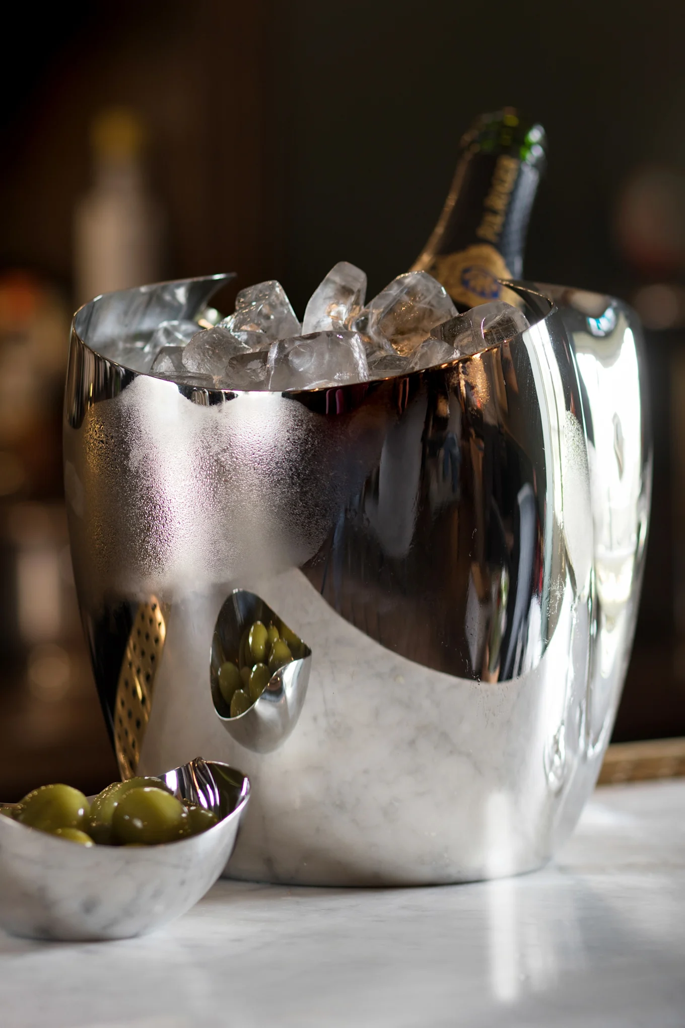 Un seau à glace argenté réfléchissant avec une bouteille de champagne et de la glace, à côté de deux petits bols d'olives vertes sur du marbre.