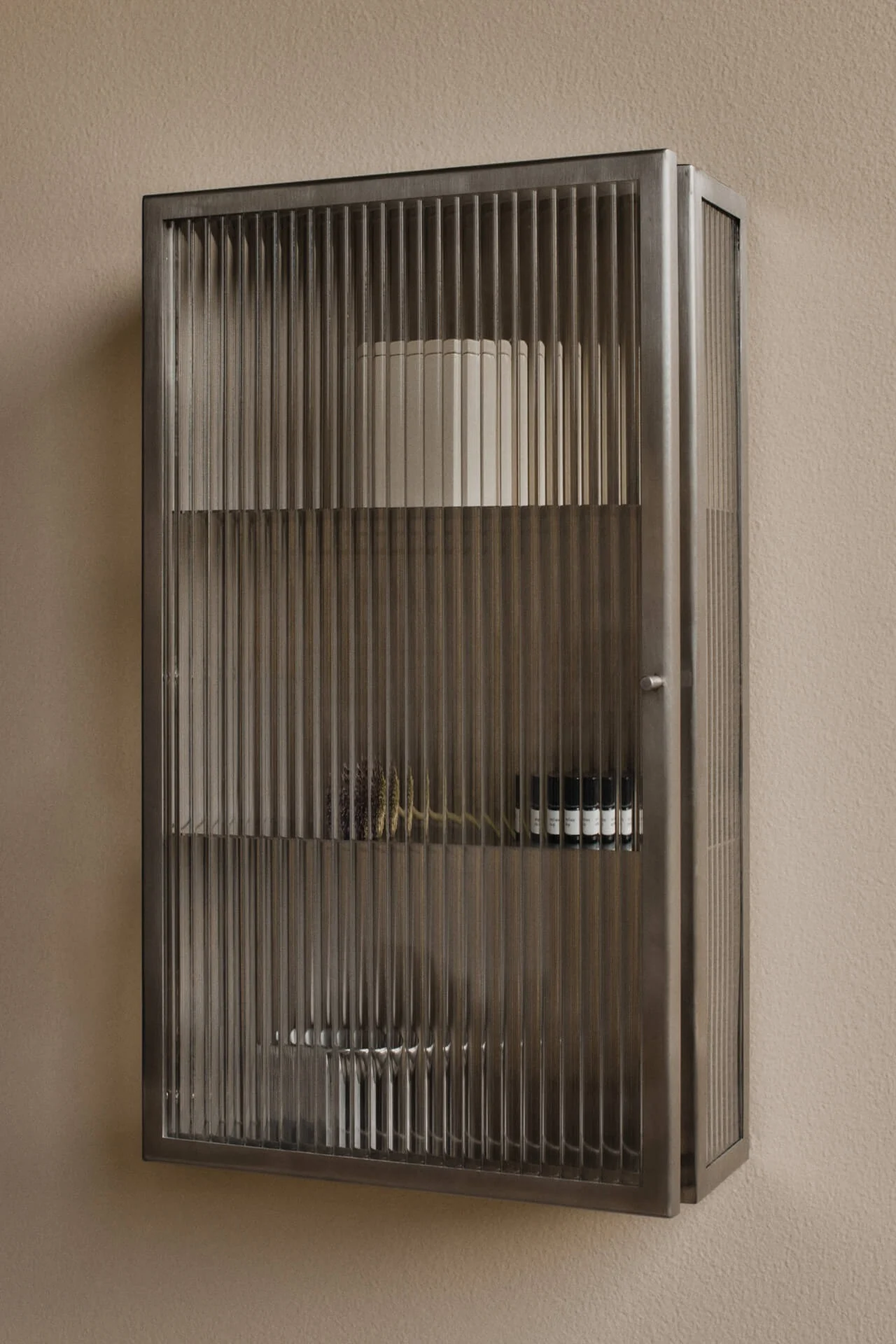 Armoire murale en métal foncé avec portes en verre cannelé sur un mur beige. Contient des étagères avec un cylindre blanc, des plantes séchées et de petites bouteilles sombres.