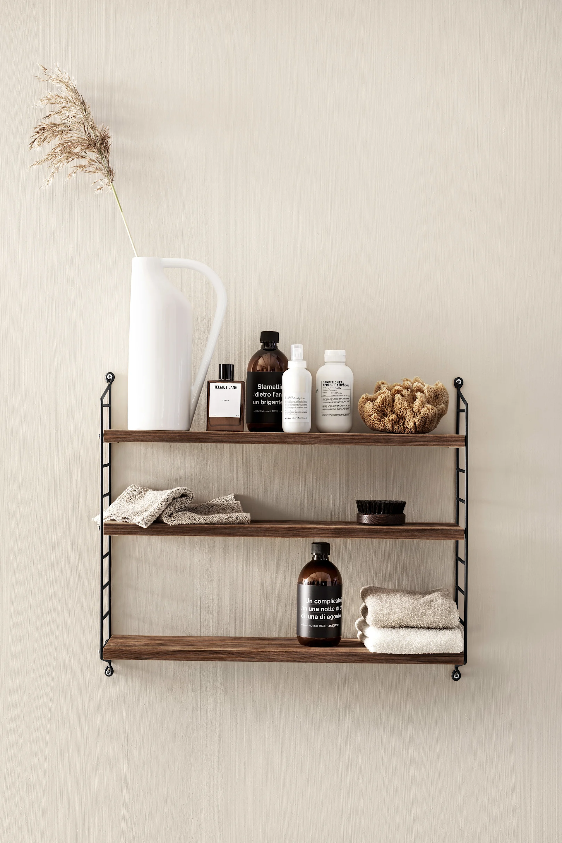 Étagère à trois niveaux en bois foncé sur mur beige avec bouteilles, serviettes, brosse et herbe de la pampa.