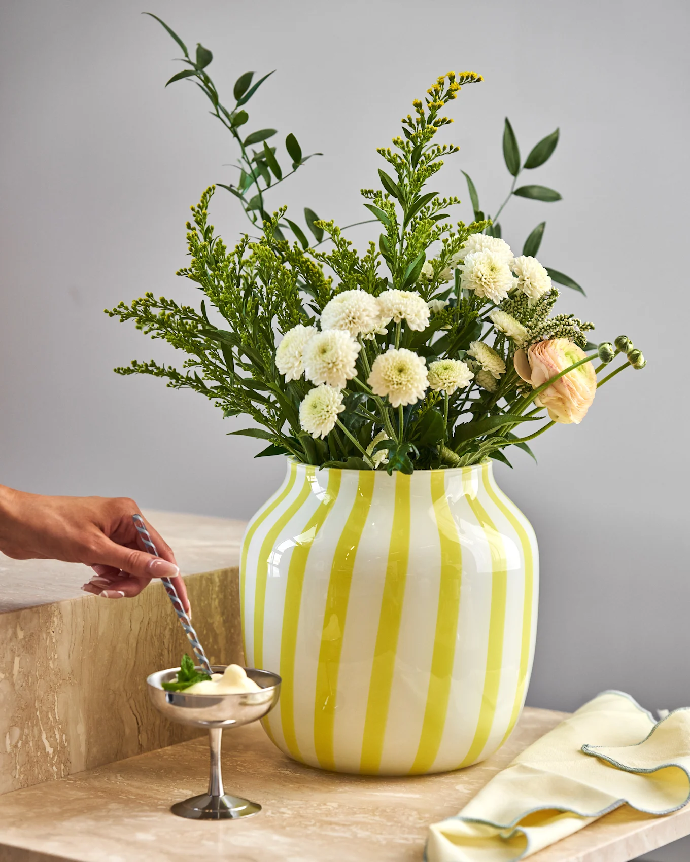 Main remue un dessert dans un bol argenté sur du marbre. Vase rayé jaune avec chrysanthèmes blancs, fleurs jaunes et renoncule rose.