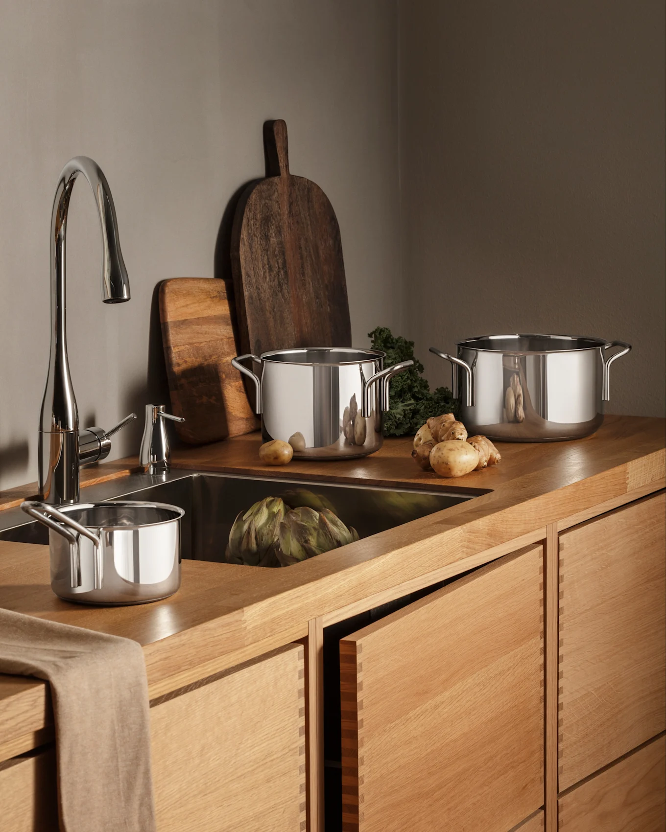 Une cuisine moderne avec des comptoirs et des armoires en bois, un robinet chromé et des casseroles en acier inoxydable, des produits frais dont artichaut et pommes de terre.
