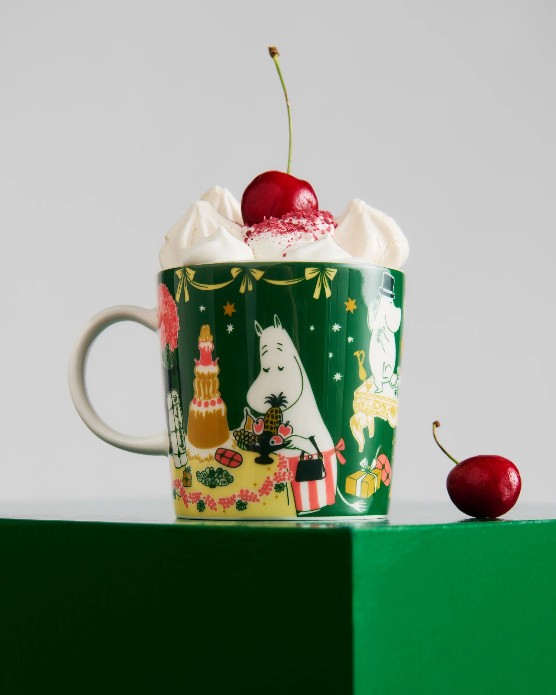 Mug vert foncé avec personnages Moomin, garni de crème fouettée, de vermicelles rouges et d'une cerise. Une autre cerise est sur la surface verte.