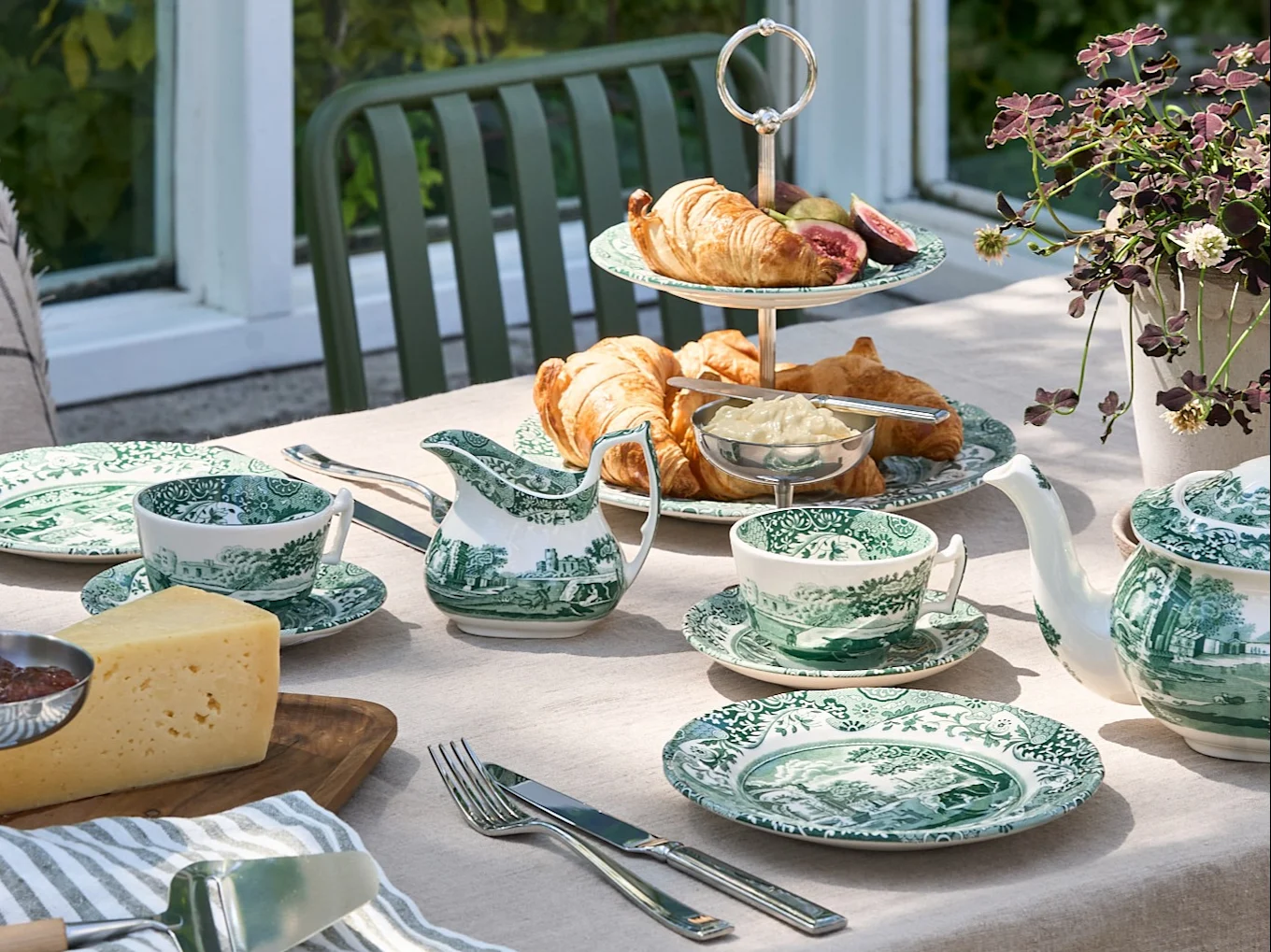 Une table d'extérieur dressée avec de la vaisselle verte et blanche, des croissants, du fromage et une théière, dans un jardin.