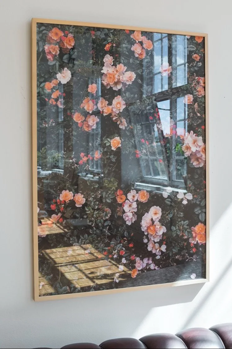 Impression encadrée d'un jardin luxuriant avec des roses pêche et des reflets de fenêtres, accrochée à un mur blanc.