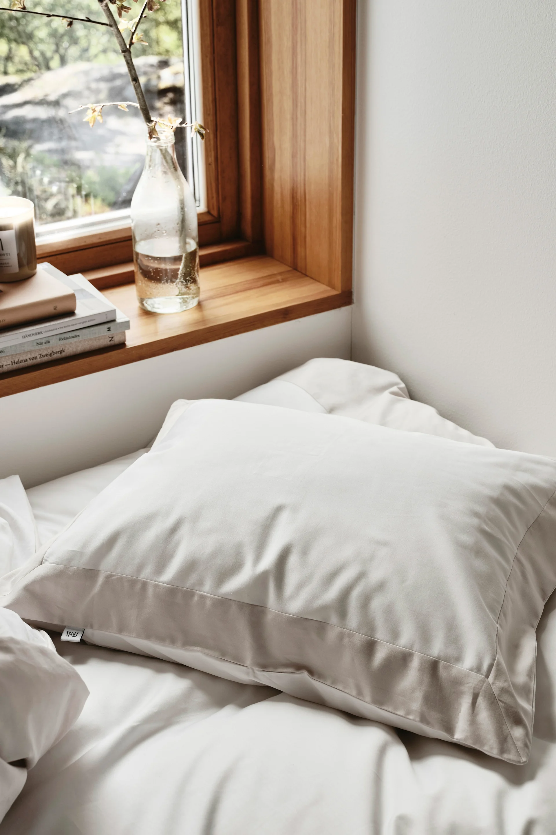 Un oreiller beige clair sur un lit blanc, à côté d'une fenêtre avec un rebord en bois, des livres et une branche de plante dans une bouteille.
