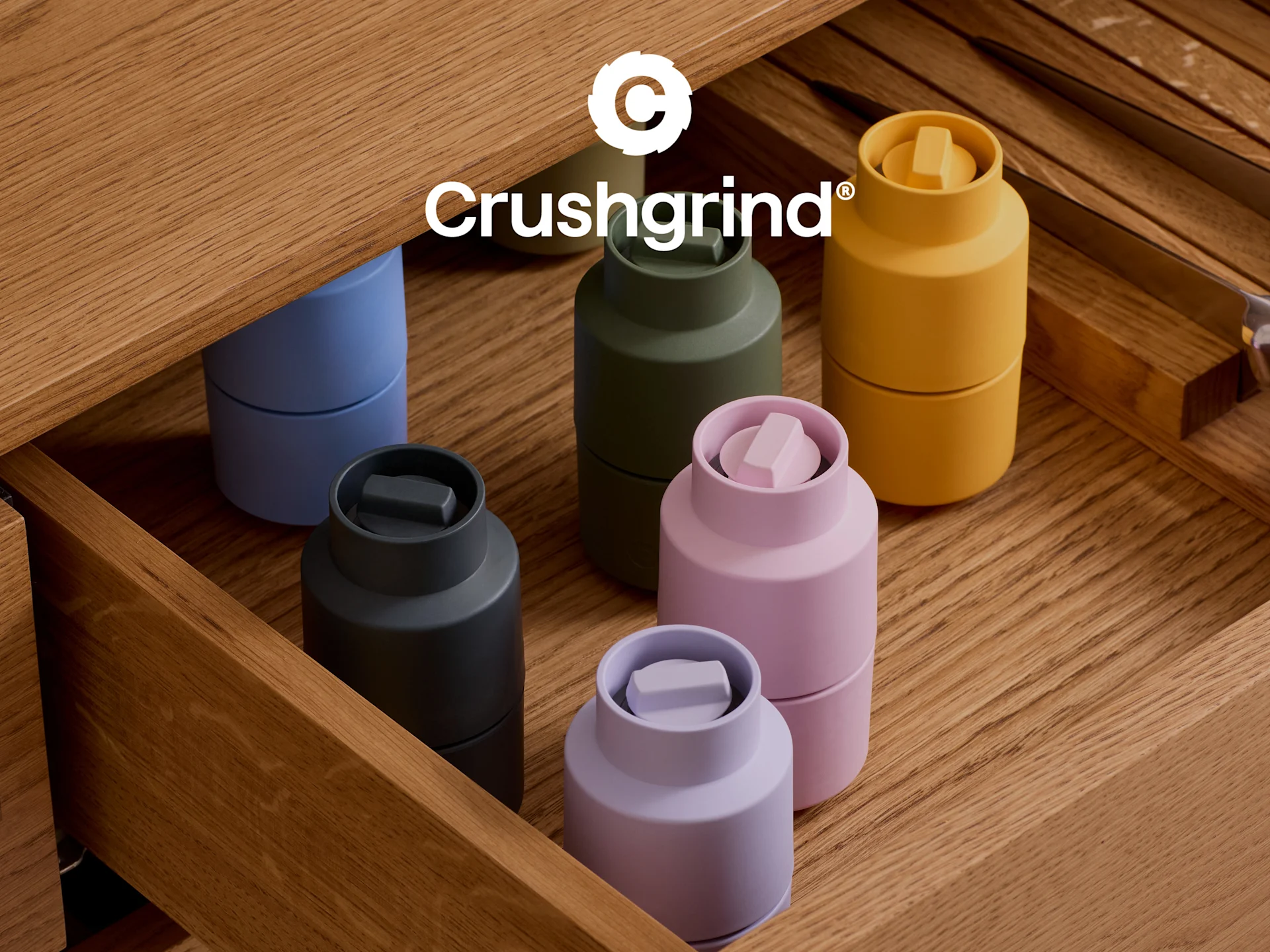 Six moulins à épices Crushgrind colorés (bleu, vert, jaune, gris, rose, violet) dans un tiroir en bois ouvert.