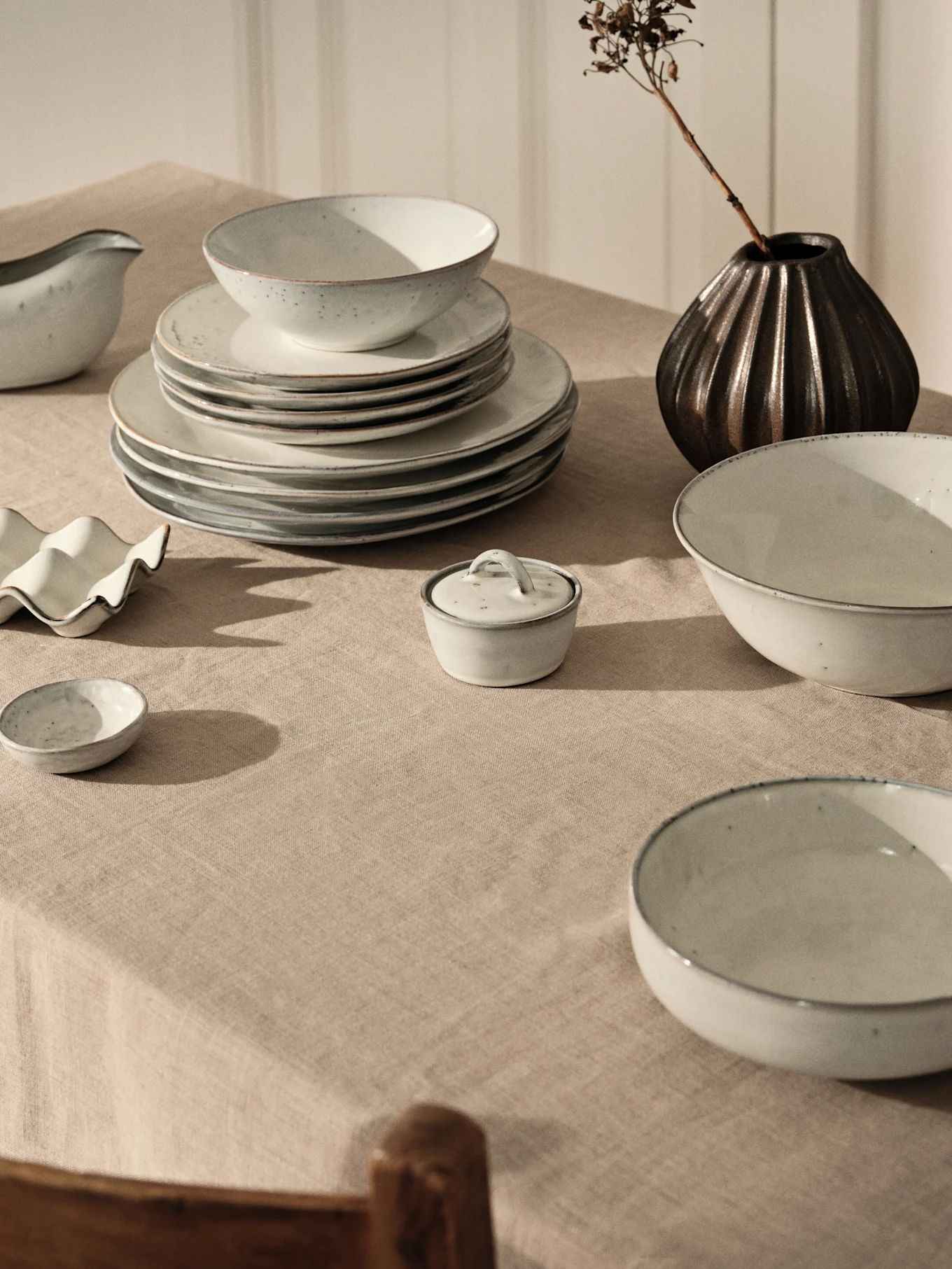 Service de vaisselle céramique gris clair avec assiettes et bols empilés sur nappe en lin beige, à côté d'un vase foncé et branche séchée.