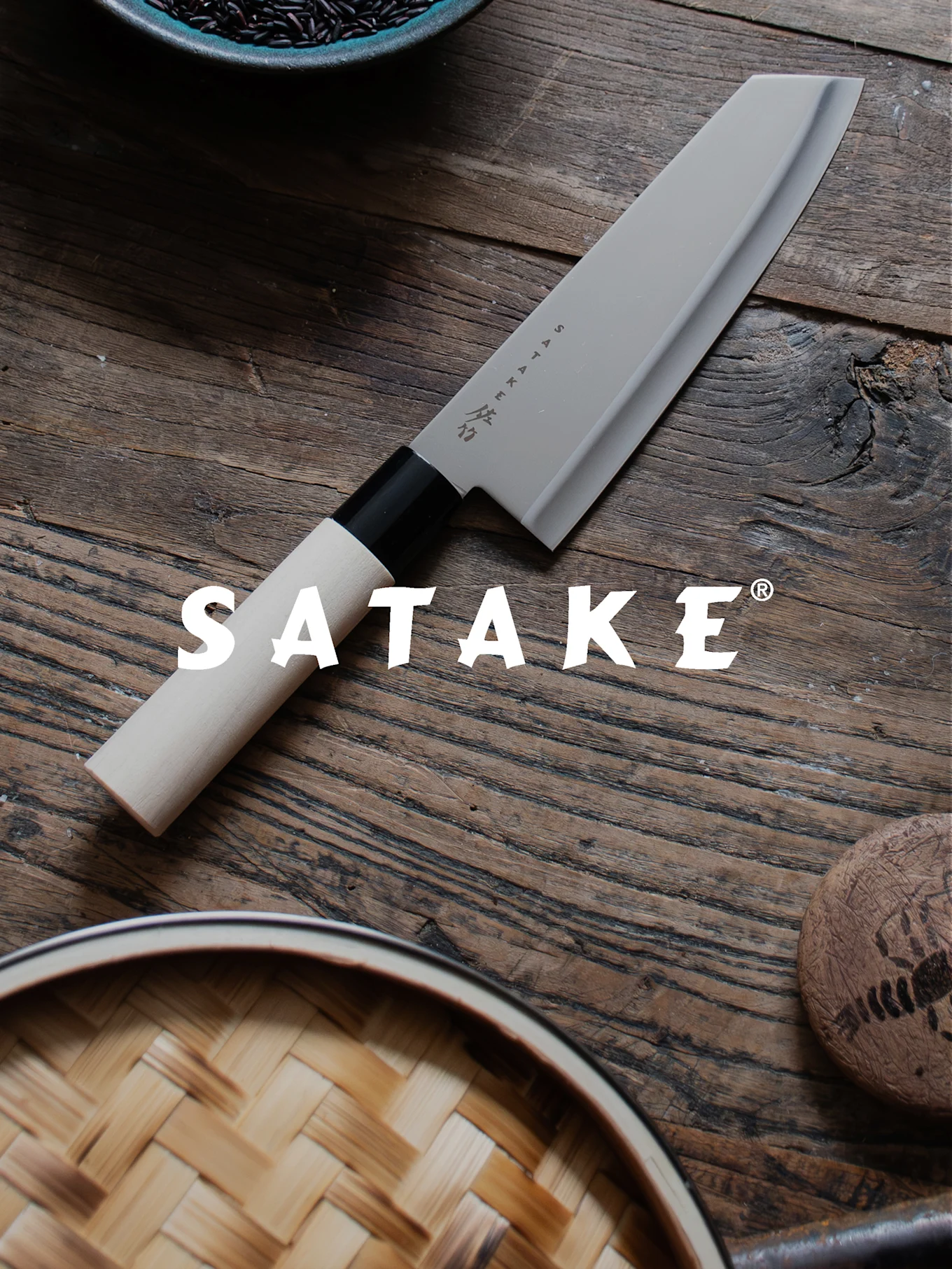 Un couteau de chef Satake avec un manche clair et une lame grise sur une table en bois, à côté de riz noir et d'un cuiseur vapeur en bambou.