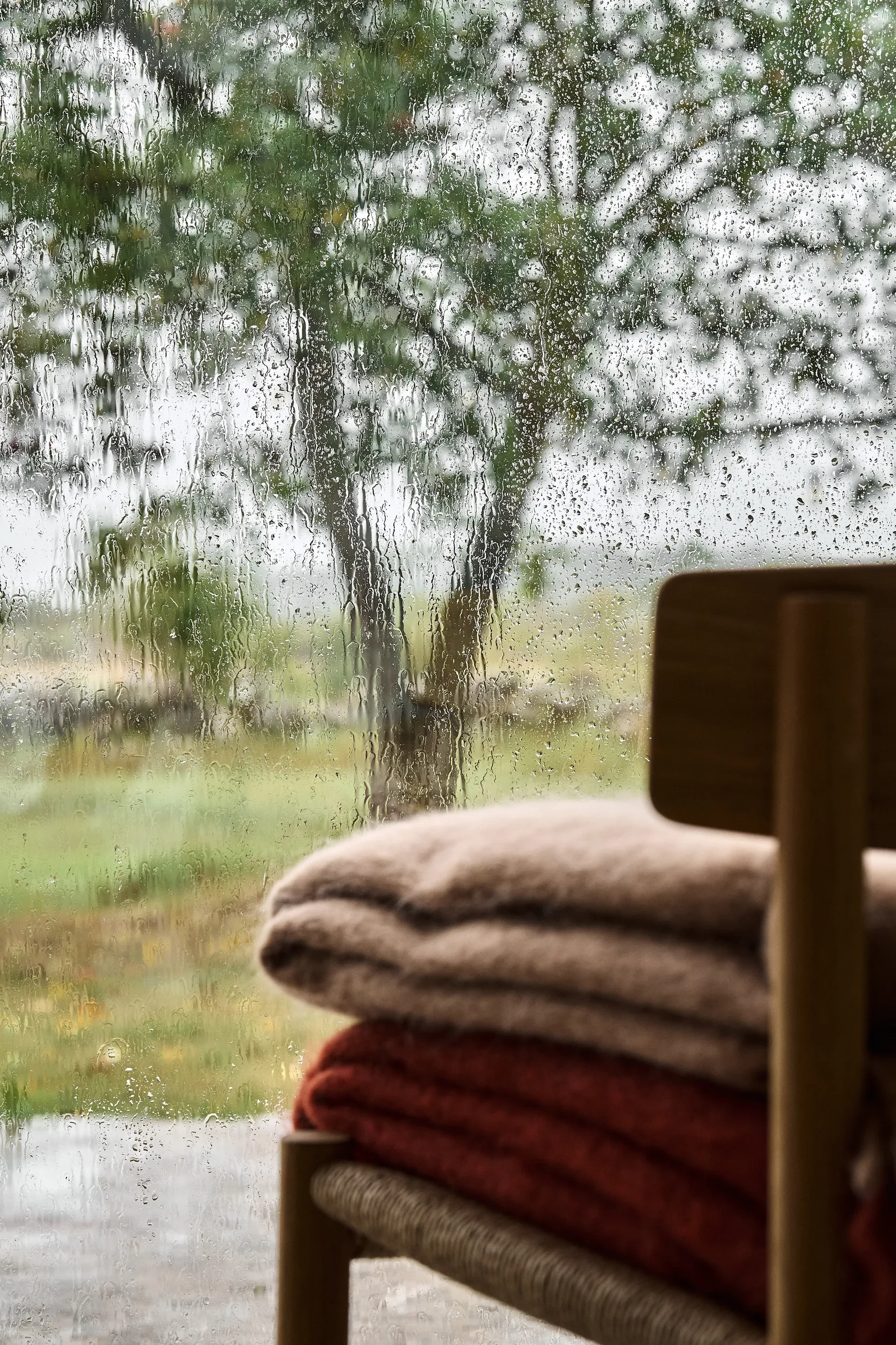 Couvertures empilées sur une chaise en bois devant une fenêtre pluvieuse avec des arbres flous.