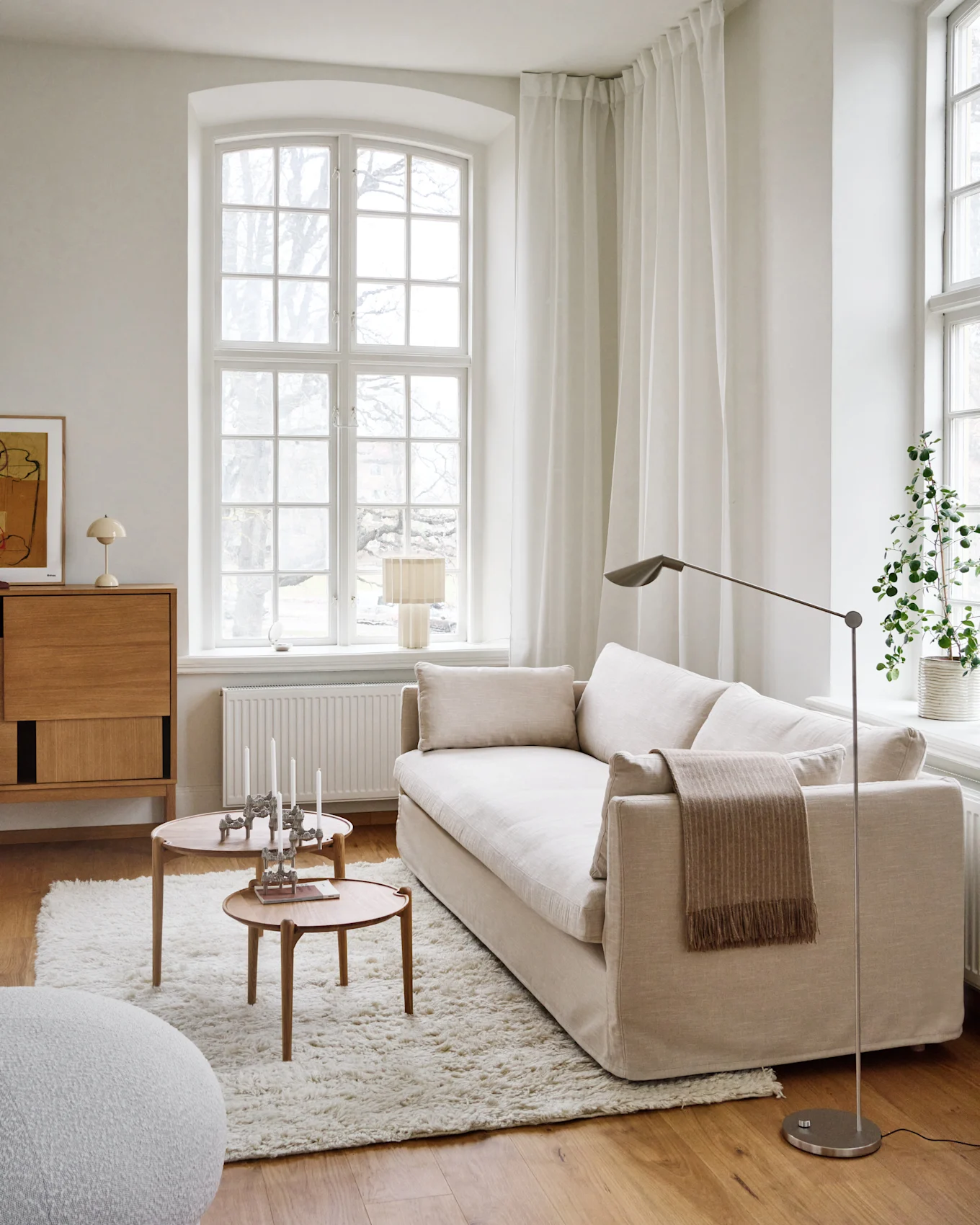 Un salon lumineux et minimaliste avec un canapé beige clair, deux tables basses rondes en bois avec des bougies, un tapis blanc à poils longs et une grande fenêtre.