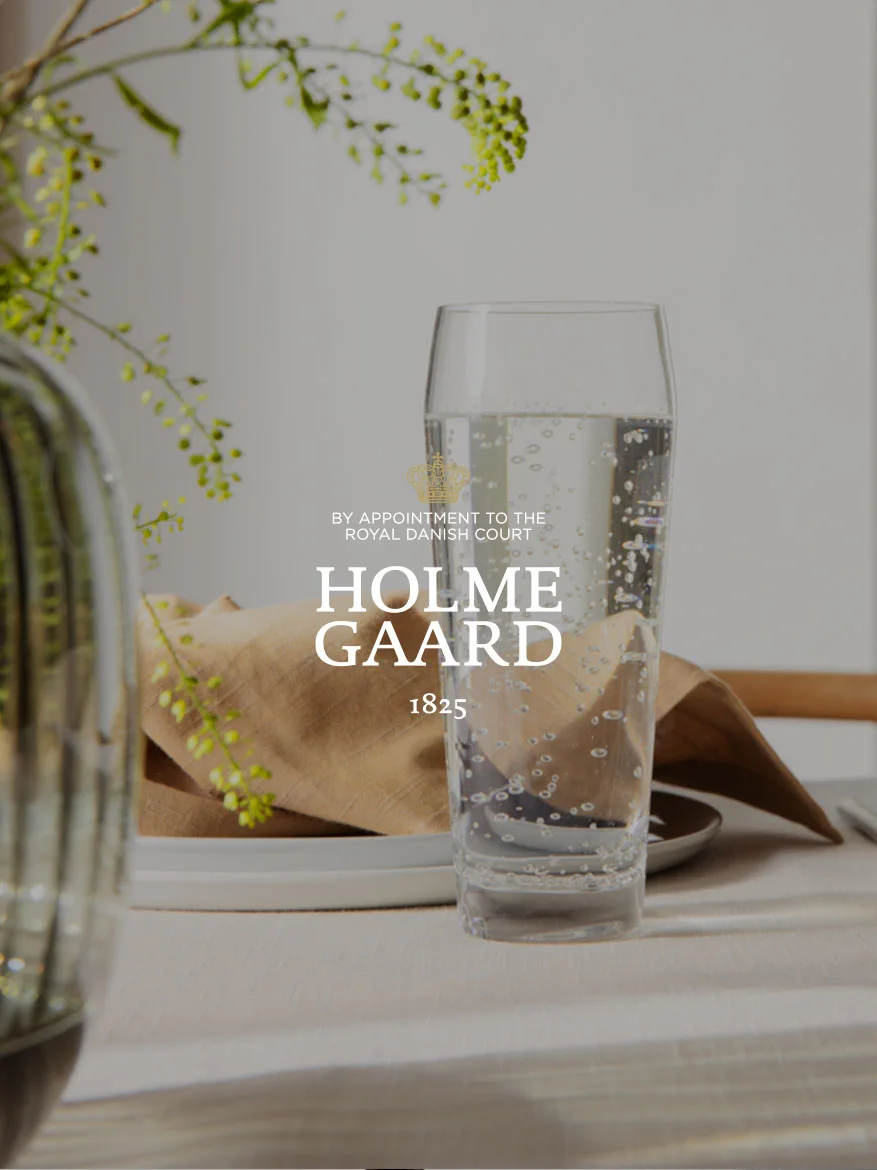 Un grand verre d'eau pétillante à côté d'une serviette marron et d'une plante, avec le logo Holmegaard superposé.