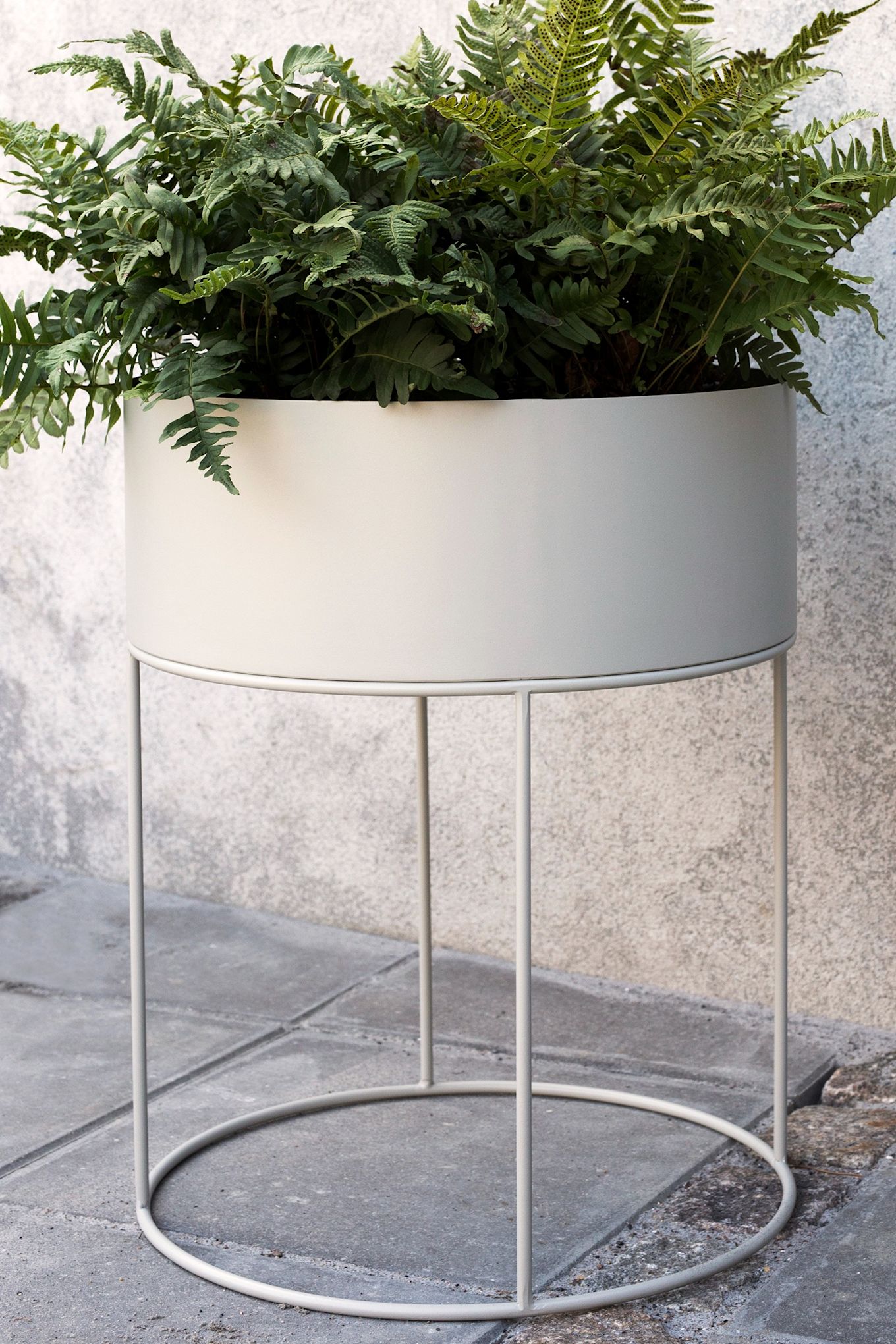 La grande jardinière ronde en métal de Ferm Living, avec une grande fougère verte, se trouve sur un patio. 