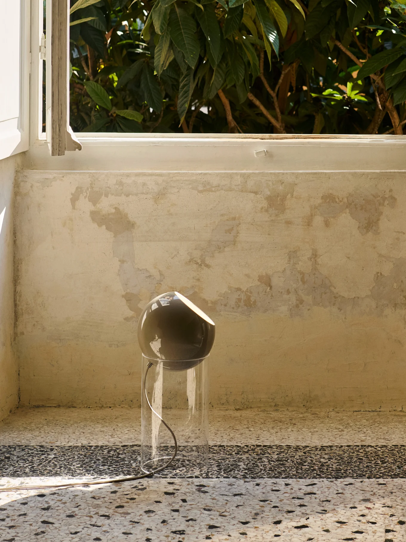 Une lampe moderne avec une tête sphérique foncée et une base cylindrique transparente repose sur un sol tacheté près d'une fenêtre avec un feuillage vert à l'extérieur.