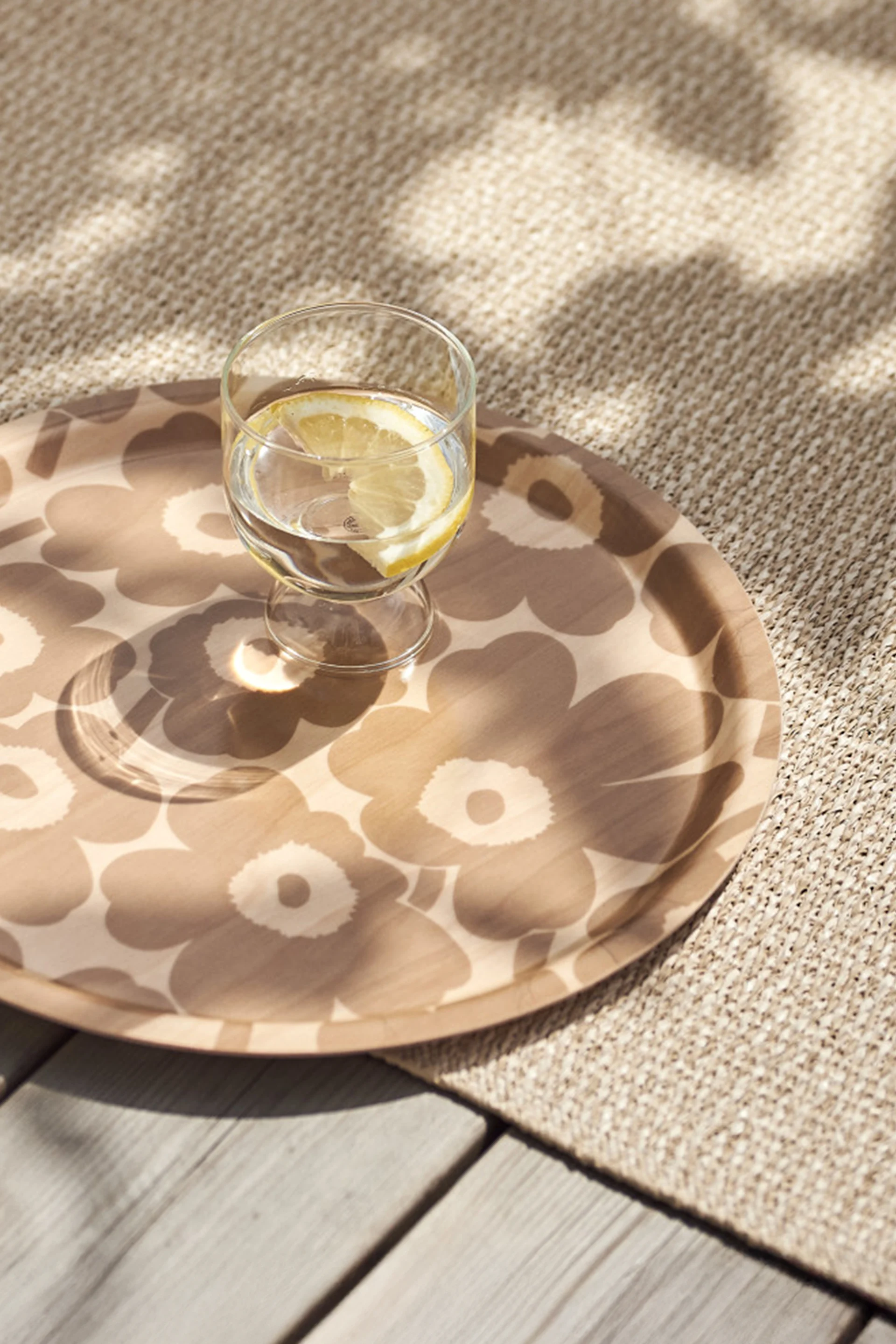 Verre d'eau avec tranche de citron sur un plateau rond à motifs, posé sur un tapis tissé beige et une terrasse en bois sous un soleil tacheté.