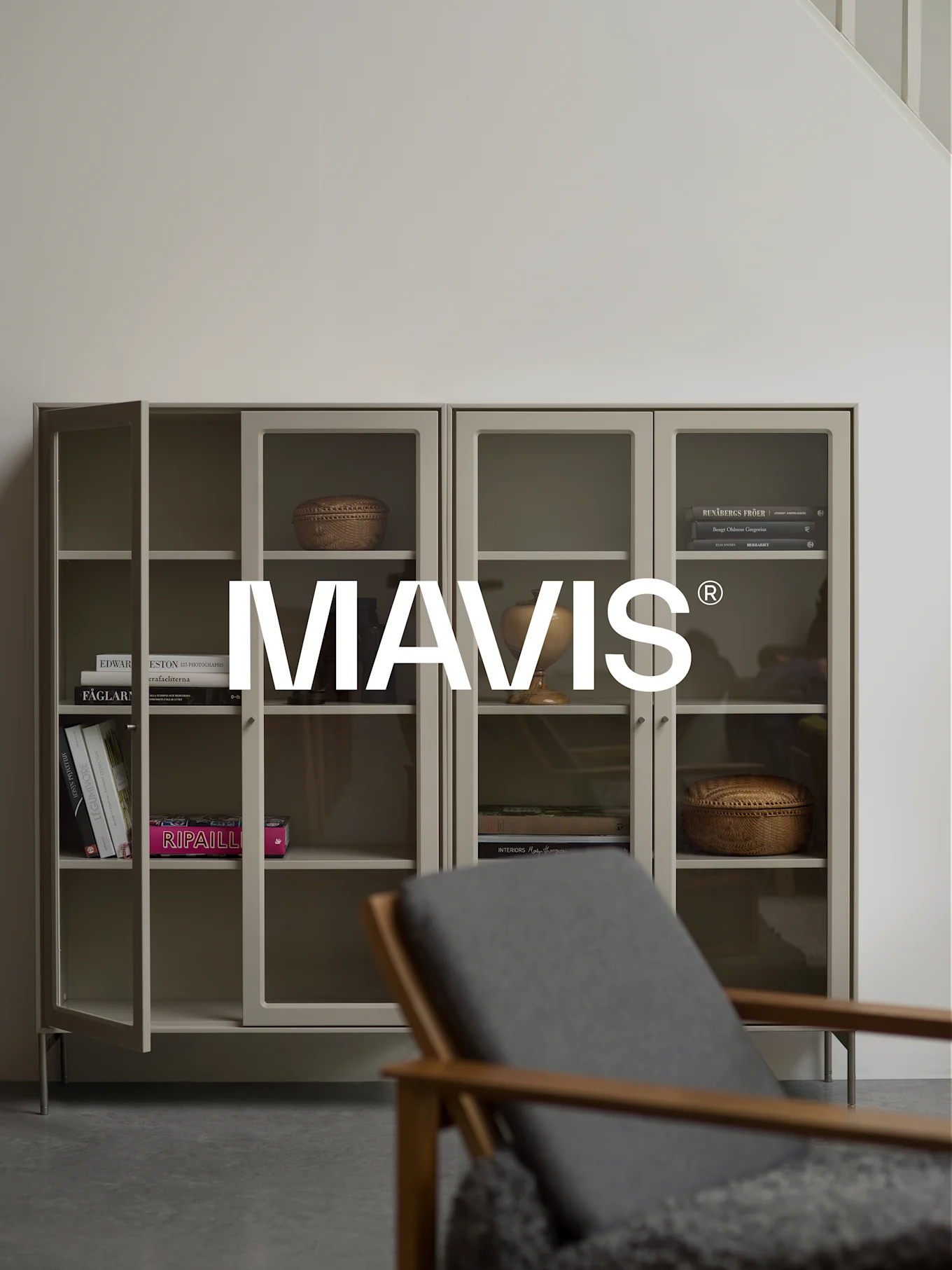 Deux armoires gris clair avec portes vitrées, présentant des livres et de la décoration. Une chaise en bois est floue au premier plan. Le logo 'MAVIS®' est centré.