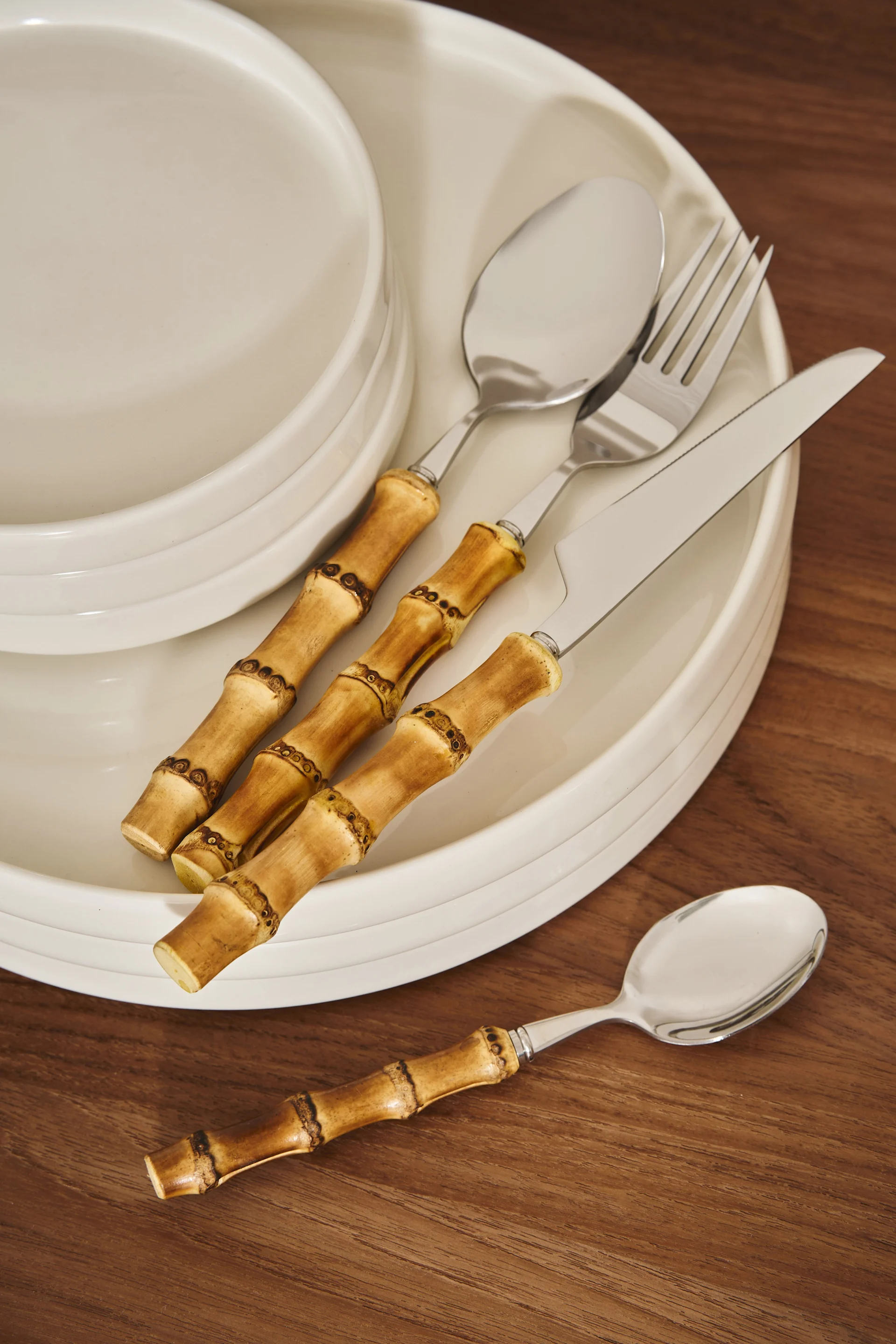 Ensemble de couverts à manche en bambou (couteau, fourchette, cuillères) posés sur des assiettes blanches et une table en bois.
