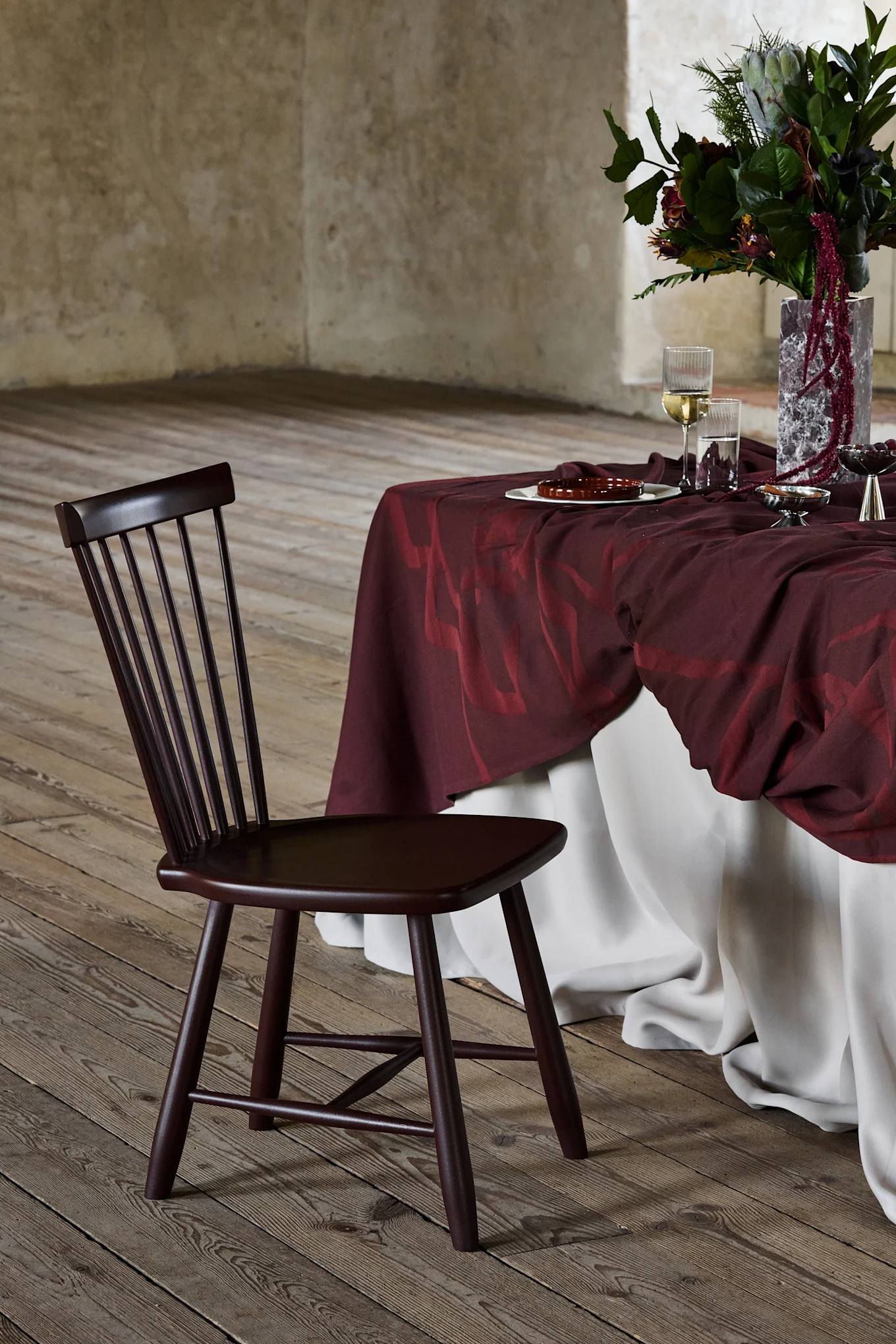 Chaise en bois marron foncé à dossier à lattes, à côté d'une table drapée de bordeaux avec un arrangement floral, sur un plancher en bois.