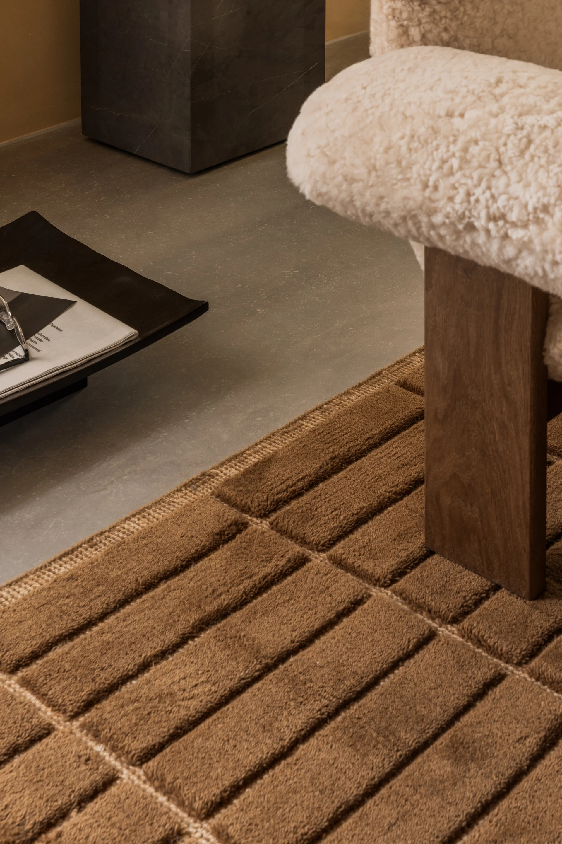 Tapis marron texturé à motifs de blocs sur un sol en béton, à côté d'une chaise crème moelleuse et d'une table d'appoint sombre.