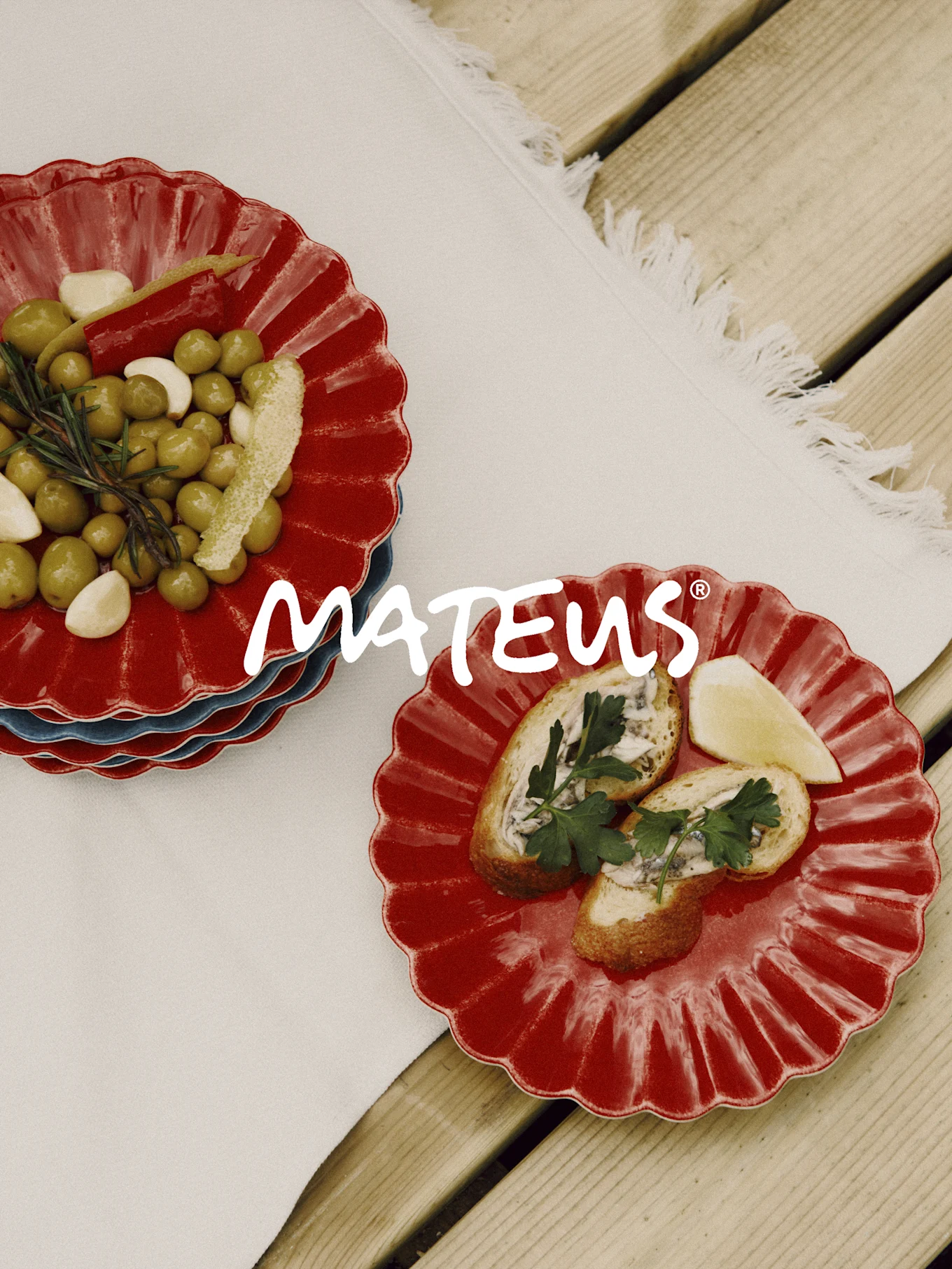 Assiettes rouges festonnées avec olives, ail, romarin et pain aux anchois sur une table en bois. Marque MATEUS.