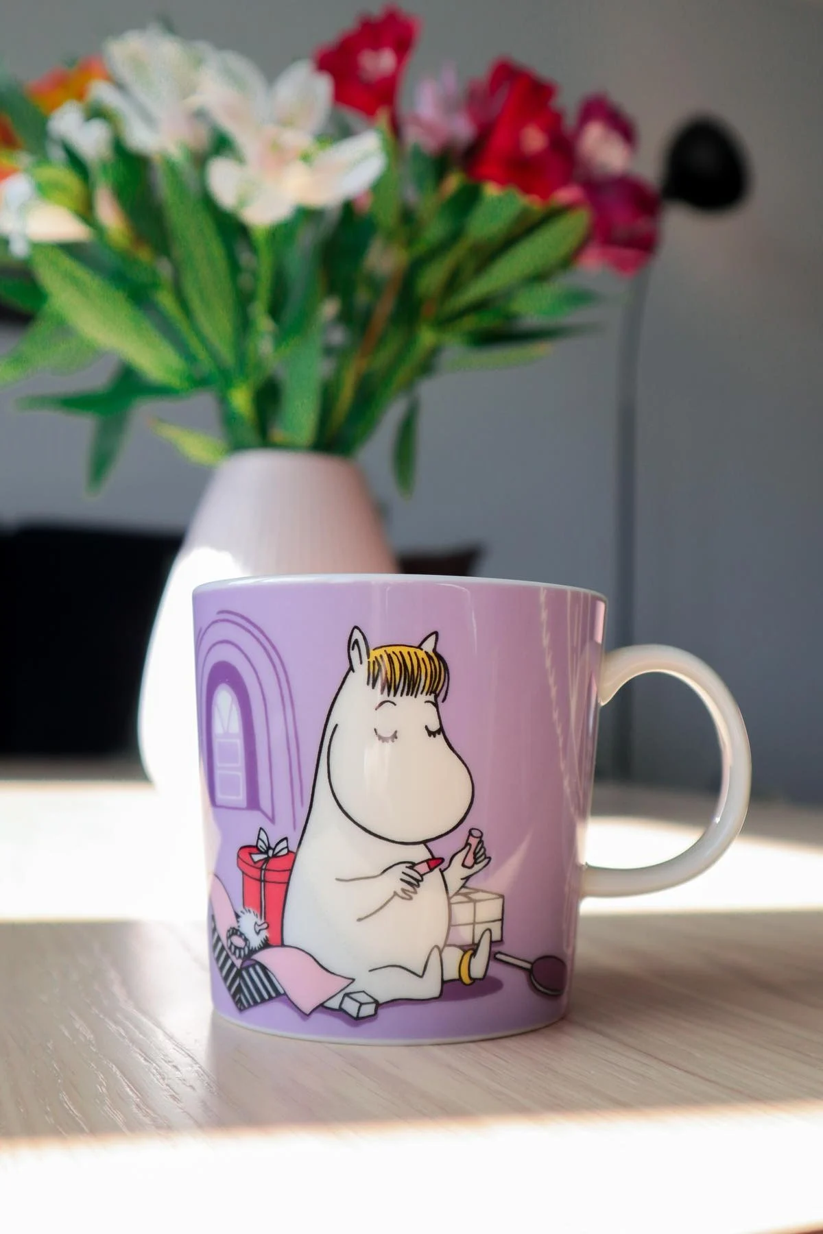 Le mug Moomin violet avec le Snorkmaiden dessus est posé sur une table, à l'arrière-plan se trouve un vase avec un bouquet de fleurs.