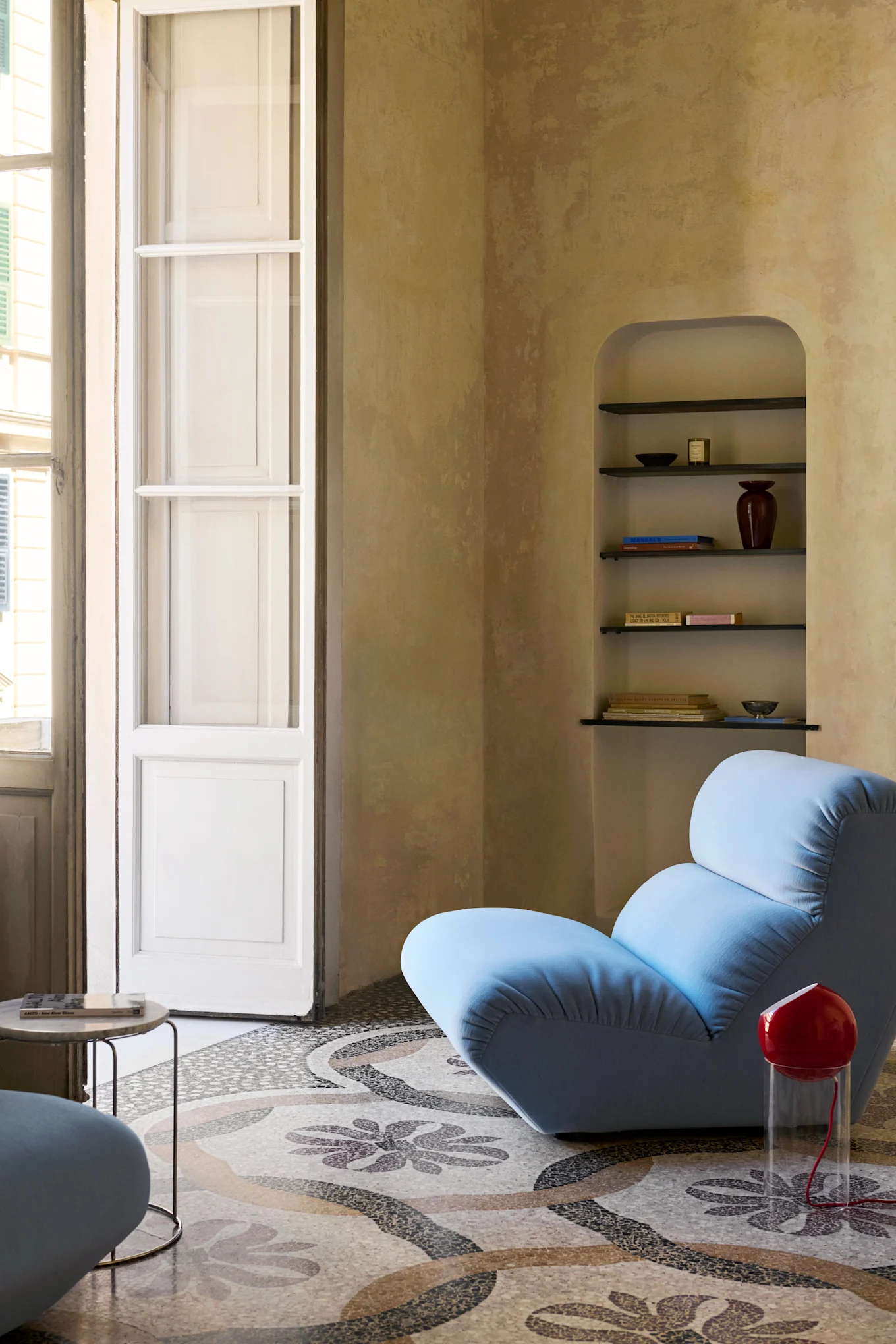 Un fauteuil lounge bleu clair sur un sol en mosaïque à motifs, à côté d'une lampe rouge, avec une étagère arquée et une haute fenêtre.