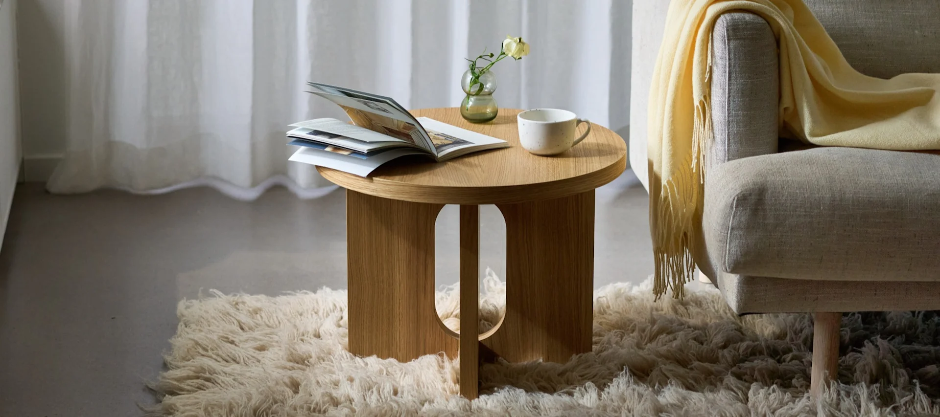 Une table d'appoint ronde en bois sur laquelle sont posés un journal, un vase et une tasse, à côté d'un fauteuil recouvert d'un plaid jaune, posé sur un tapis duveteux.