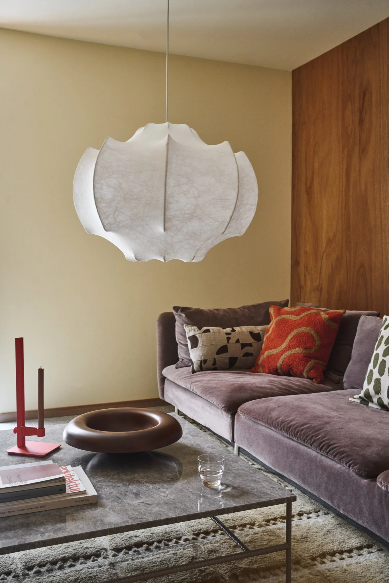 Un salon avec une lampe en papier blanc ondulé, un canapé en velours couleur prune, une table en marbre gris et divers éléments de décoration.