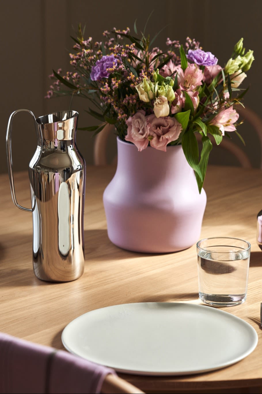 Vous voyez ici le vase Dorotea violet avec un bouquet de fleurs roses et violet posé sur une table à côté de la carafe Dorotea argentée.