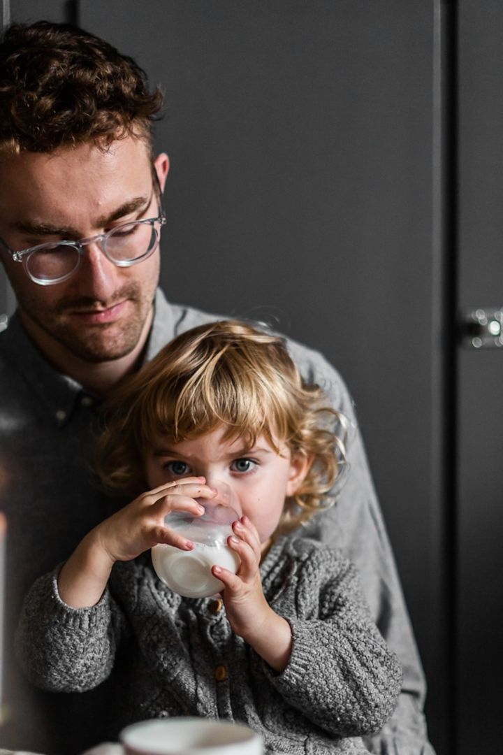 Le temps passé ensemble est certainement le cadeau le plus précieux pour la fête des pères. Ici, vous voyez un petit enfant assis sur les genoux de son père et buvant dans un verre de Scandi Living.