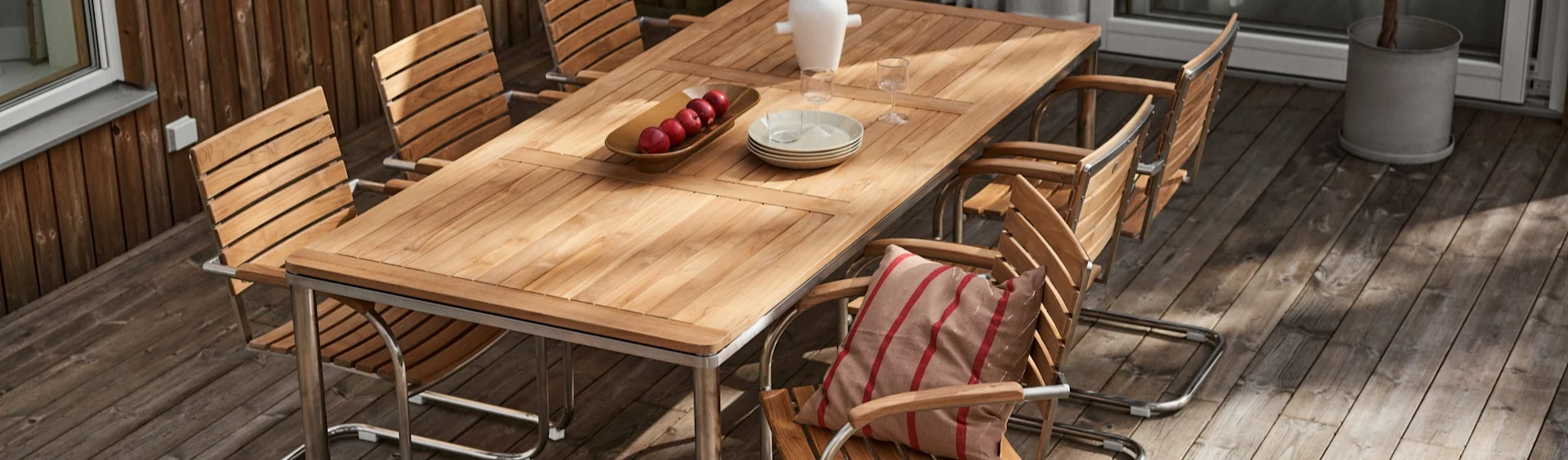 Table à manger en bois d'extérieur avec plusieurs chaises sur une terrasse. La table a des pommes, des assiettes et des verres. Une chaise a un coussin rayé.