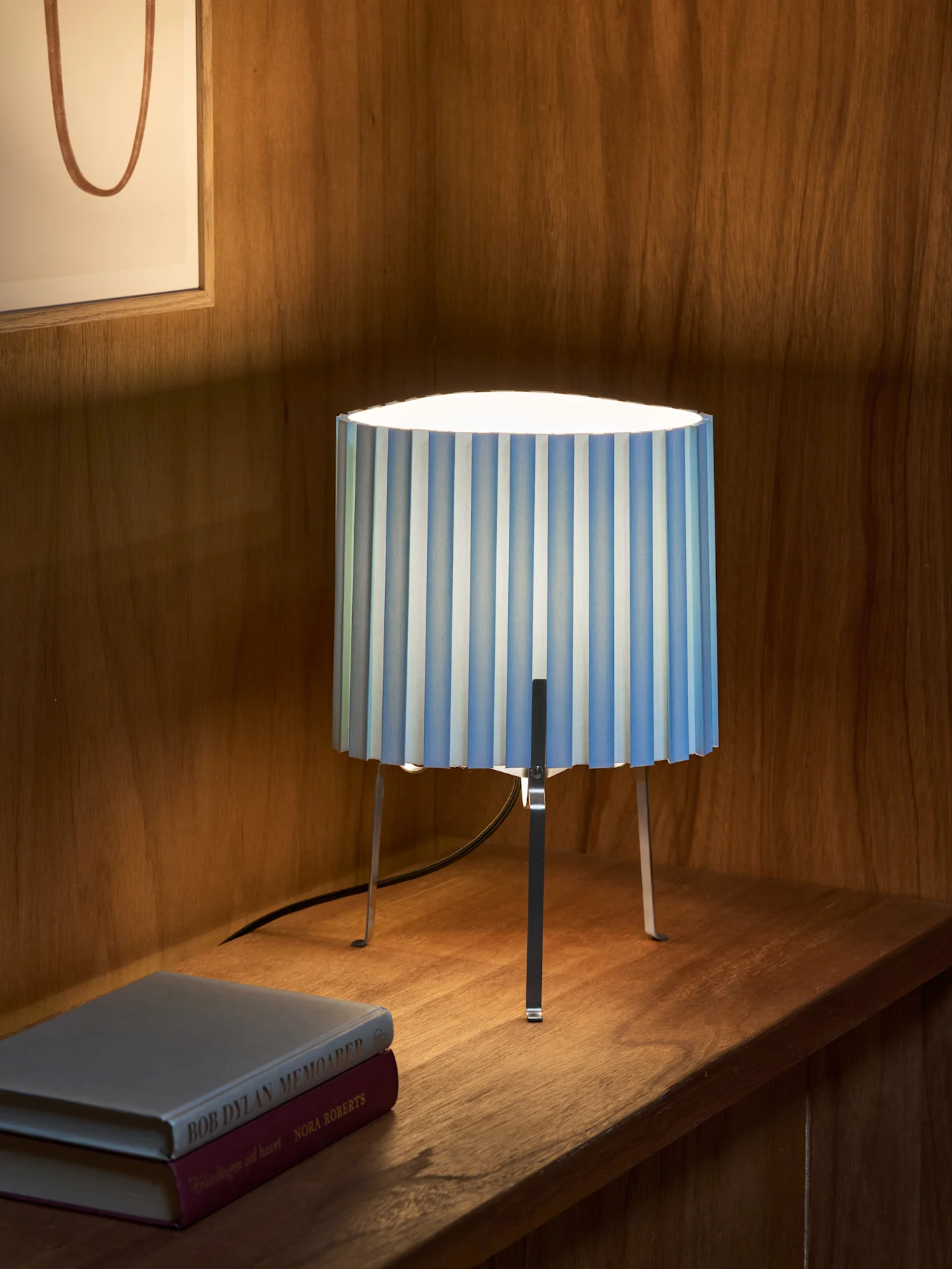 Lampe de table plissée à rayures bleues et blanches avec trois pieds métalliques sur une étagère en bois près de deux livres. Œuvre encadrée en arrière-plan.