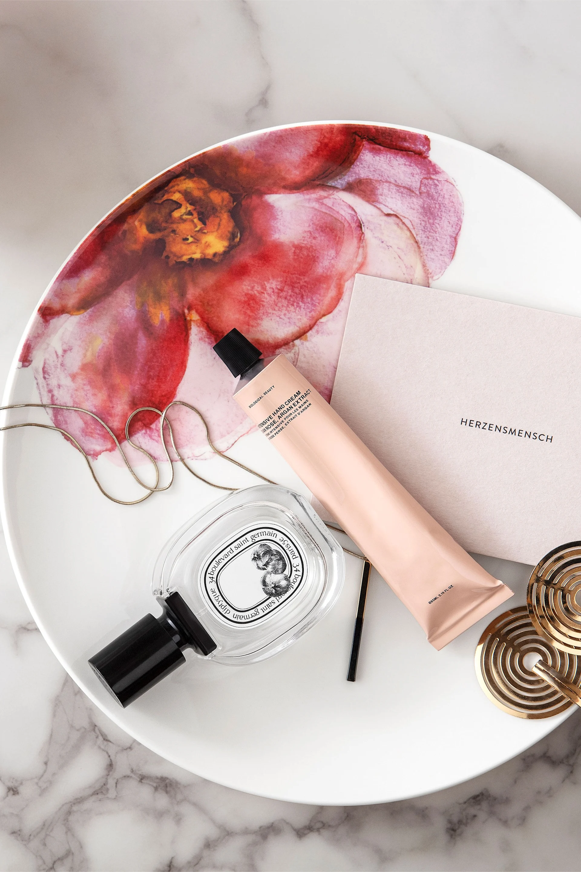 Parfum, crème mains pêche, chaîne en or et sous-verres disposés sur un plateau blanc à motif floral rose.