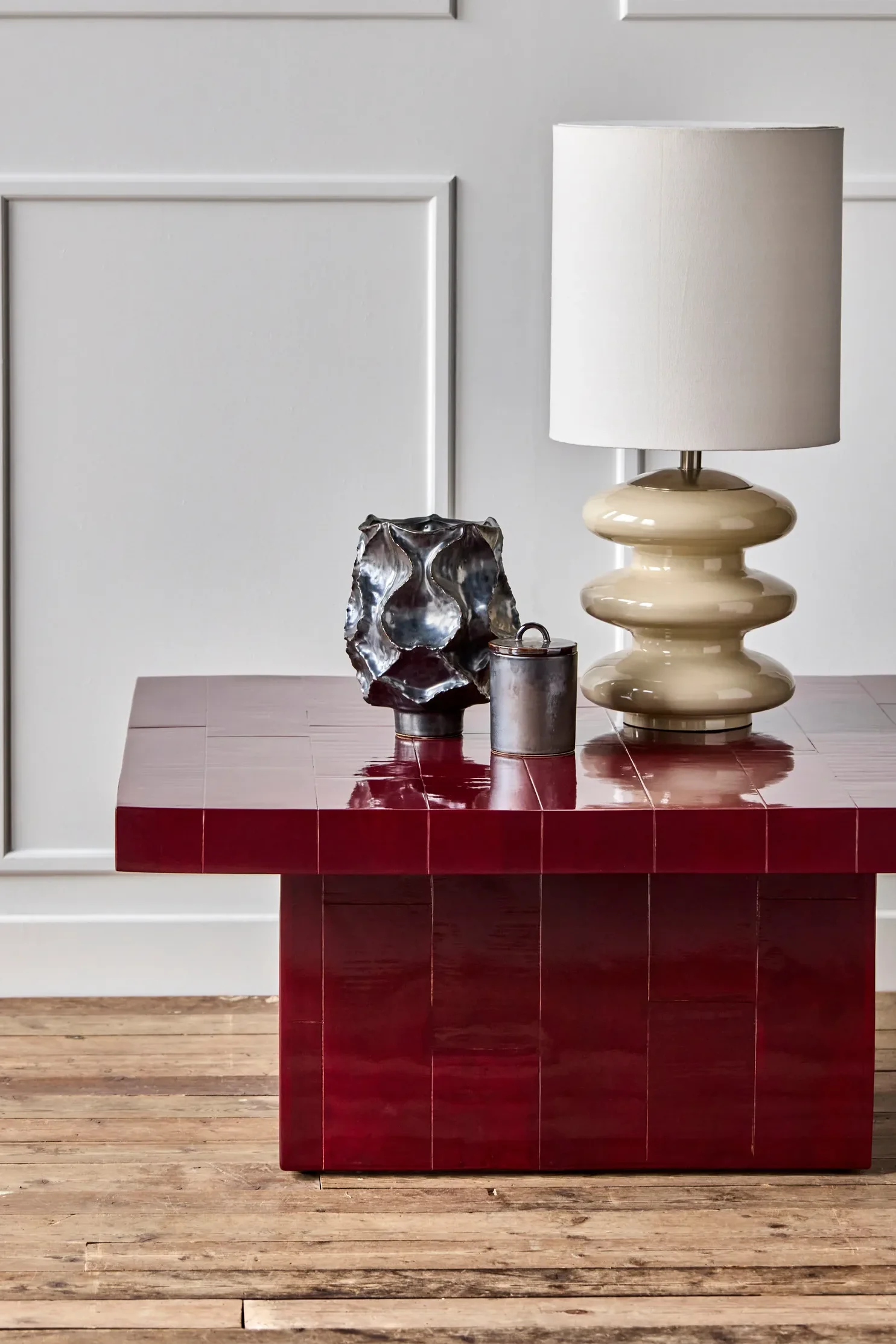 Une table basse en carreaux rouge foncé brillant, sur laquelle sont posés une lampe sculpturale de couleur crème, un vase abstrait en métal foncé et un petit récipient cylindrique.