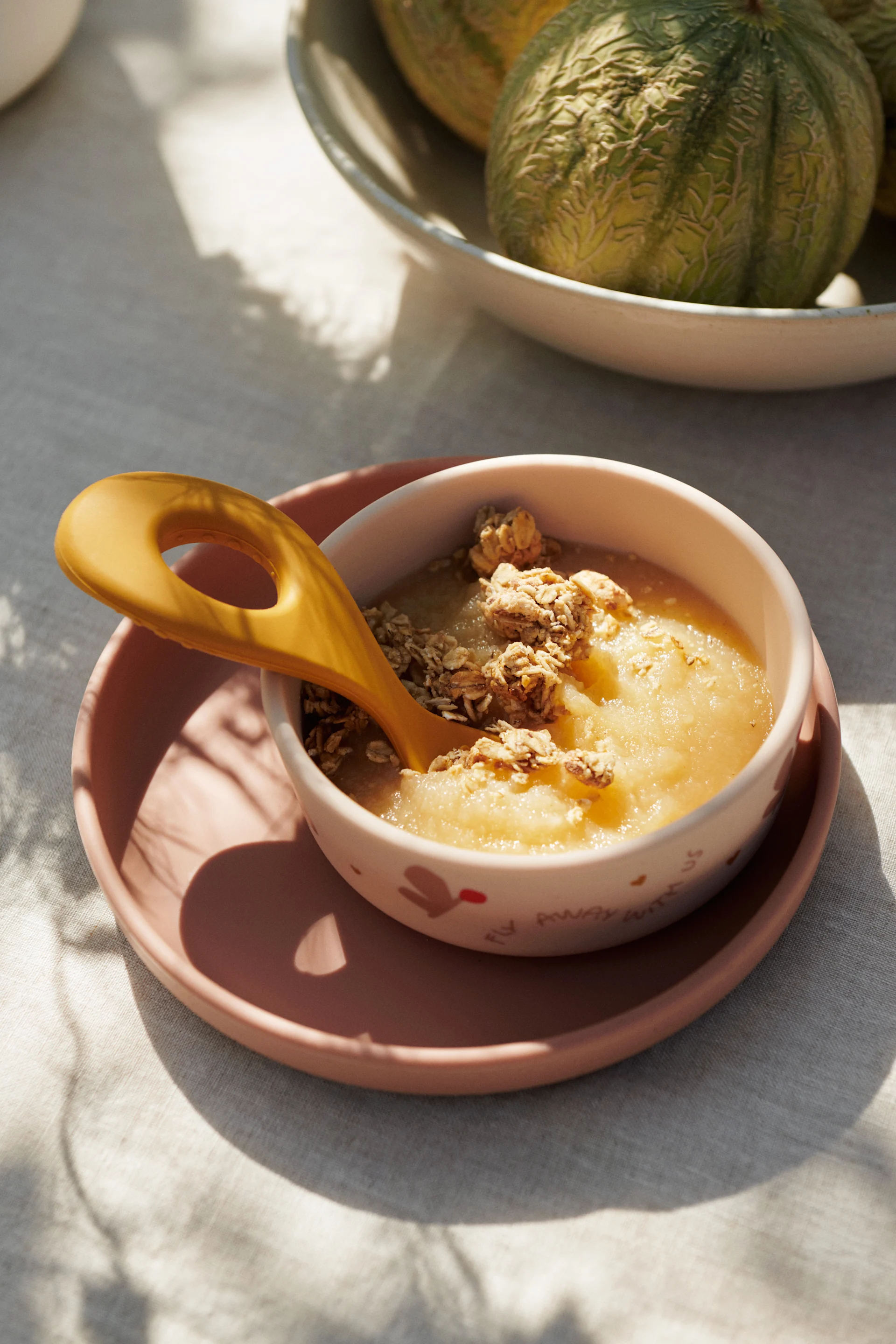 Compote de pommes avec granola et une cuillère jaune dans un bol sur une assiette rose, avec des melons.