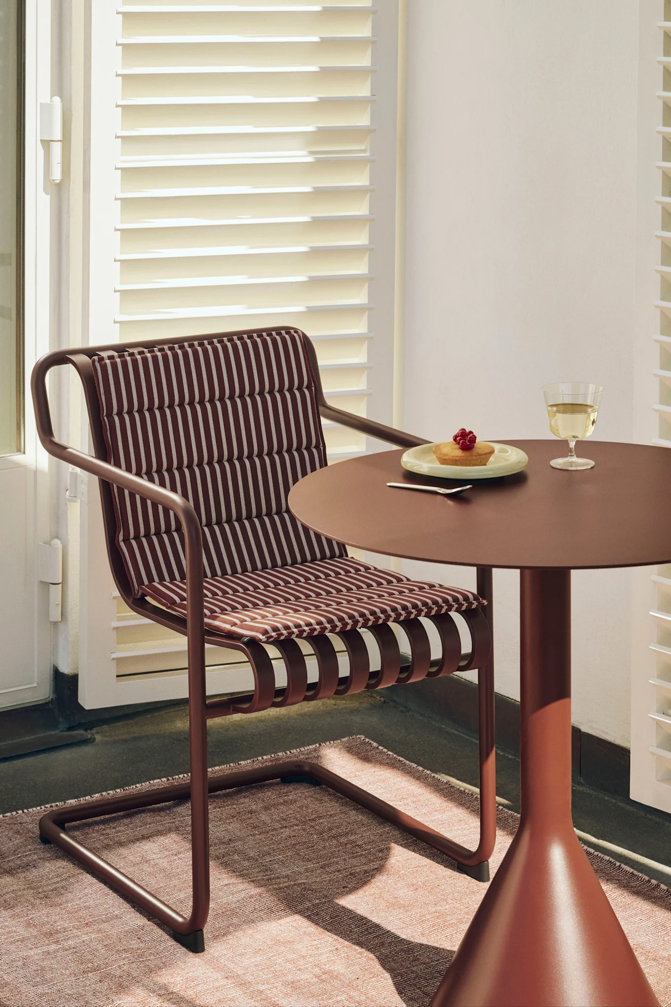Une chaise brun-rouge avec un coussin rayé à côté d'une table ronde assortie avec un gâteau et un verre de vin blanc.