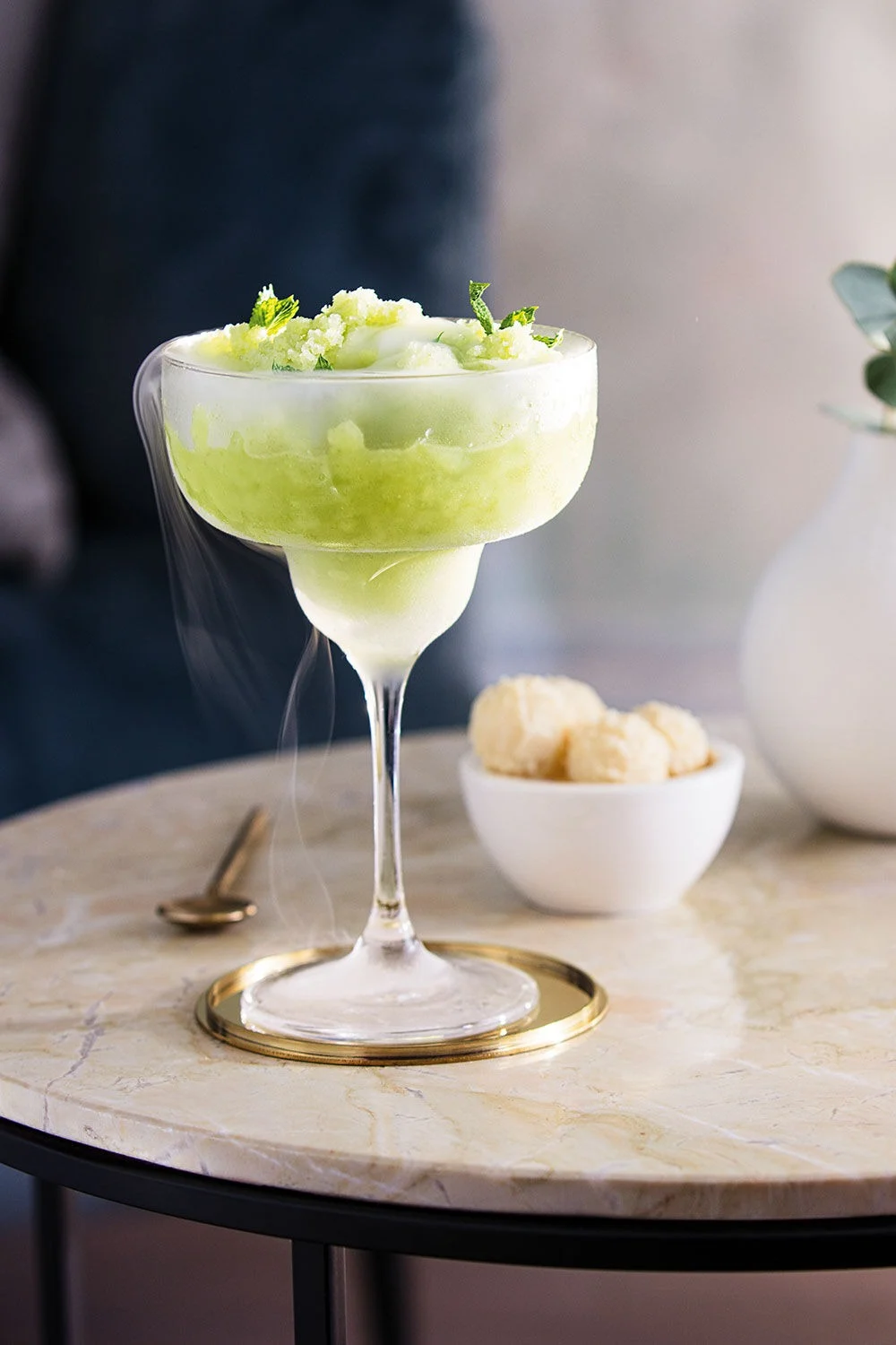 Boisson verte glacée dans un verre à margarita garnie de menthe, sur une table en marbre avec un sous-verre doré.