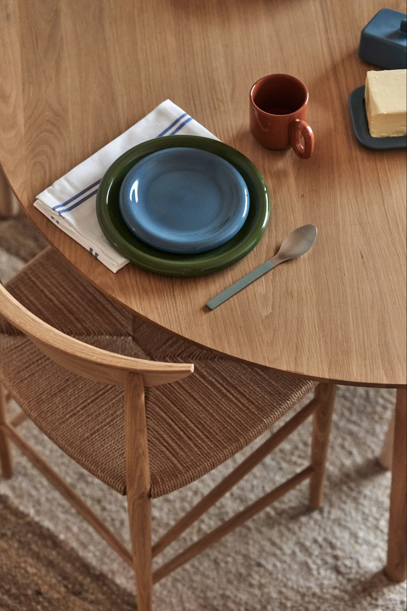 Une table en bois dressée pour une personne, avec des assiettes vertes et bleues, une serviette rayée, une tasse, une cuillère et du beurre. On aperçoit une chaise tressée.