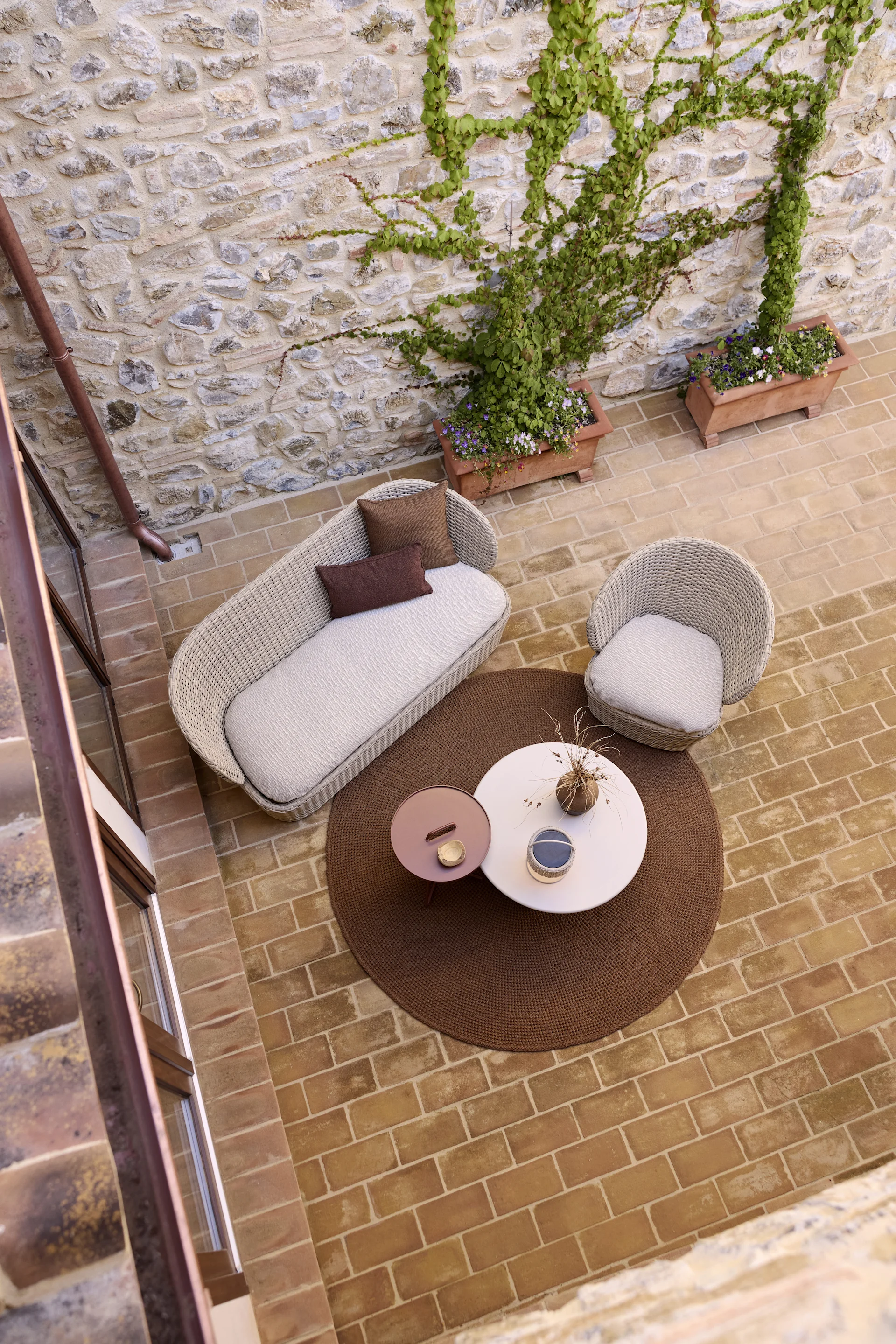 Vue plongeante d'un patio avec un canapé tressé, un fauteuil, deux tables basses rondes sur un tapis marron et des murs en pierre recouverts de lierre.