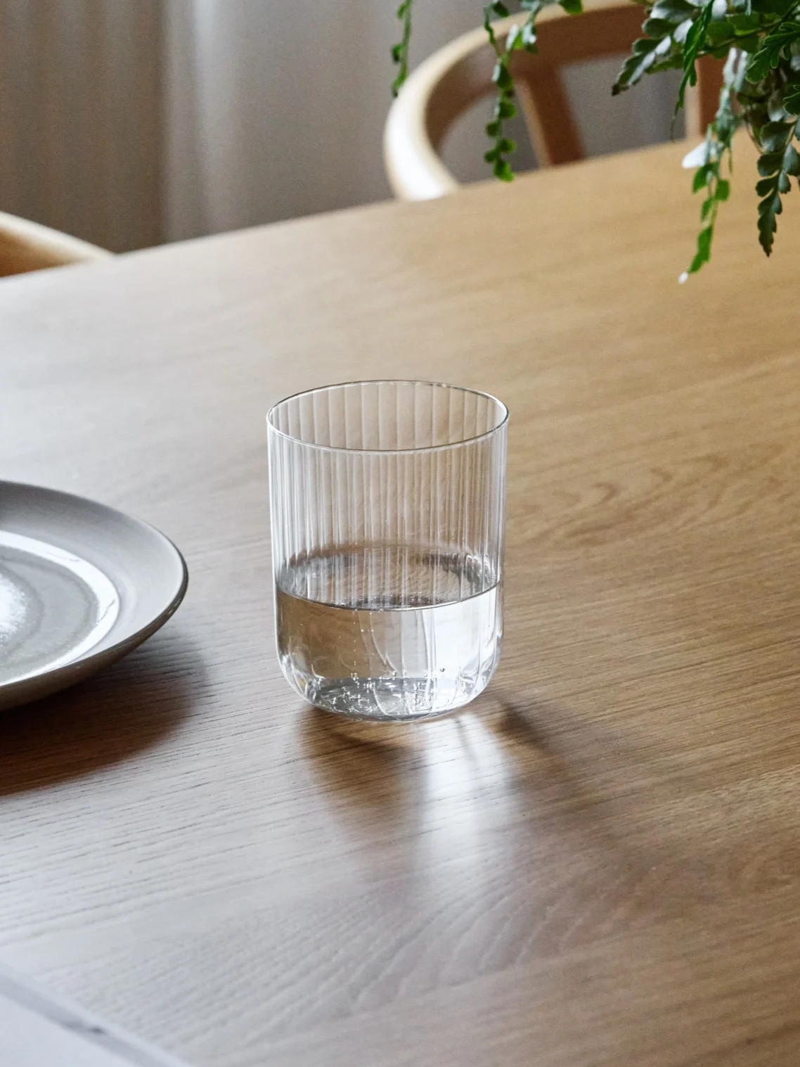 Un verre strié, à moitié rempli d'eau, posé sur une table en bois à côté d'une assiette en céramique.