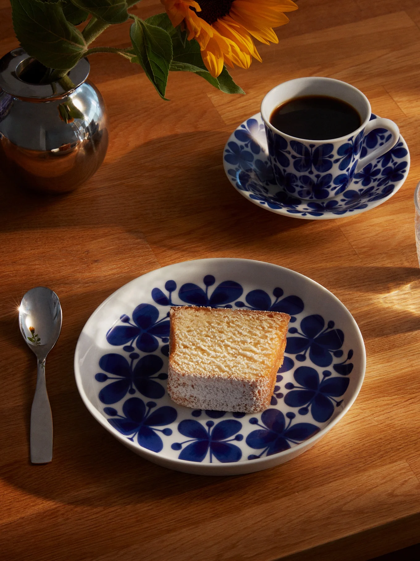 Une tranche de gâteau sur une assiette florale bleue et blanche, avec une tasse de café, un verre d'eau, une cuillère et un tournesol dans un vase argenté sur une table en bois ensoleillée.