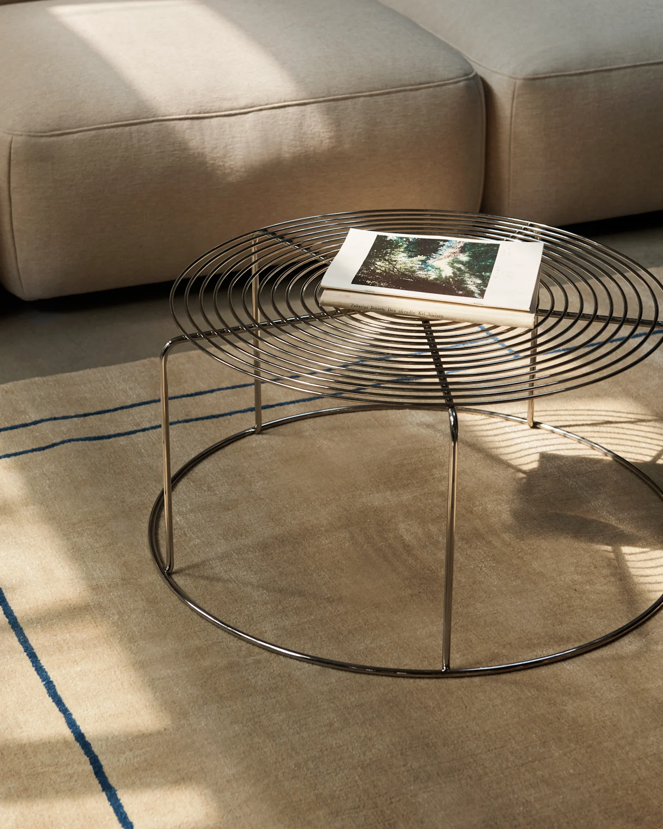 Table basse ronde en métal avec un livre sur un tapis beige, à côté d'un canapé.