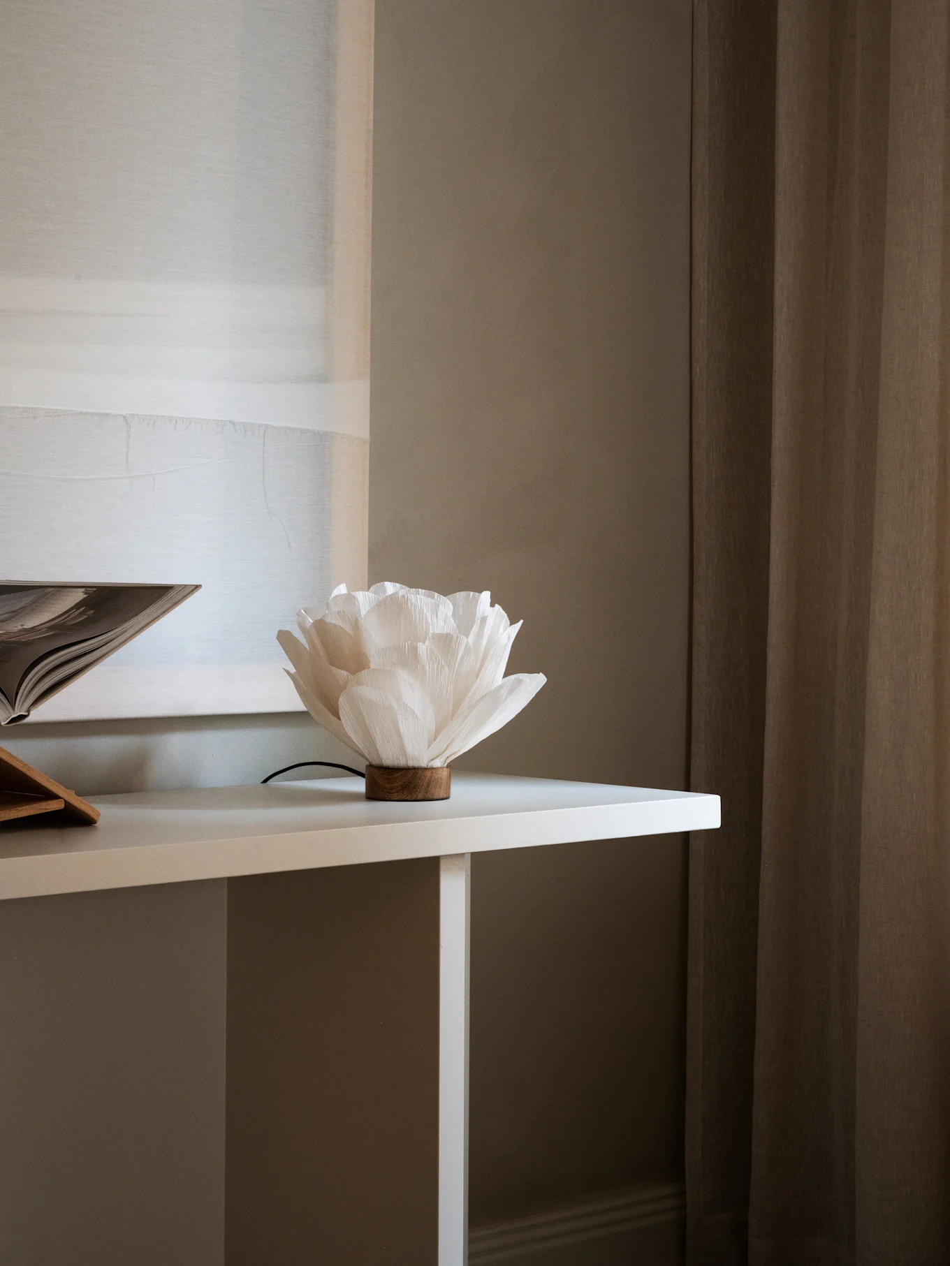 Lampe en papier blanche en forme de fleur avec une base en bois sur une table blanche, à côté d'un livre ouvert. Rideaux marrons en arrière-plan.