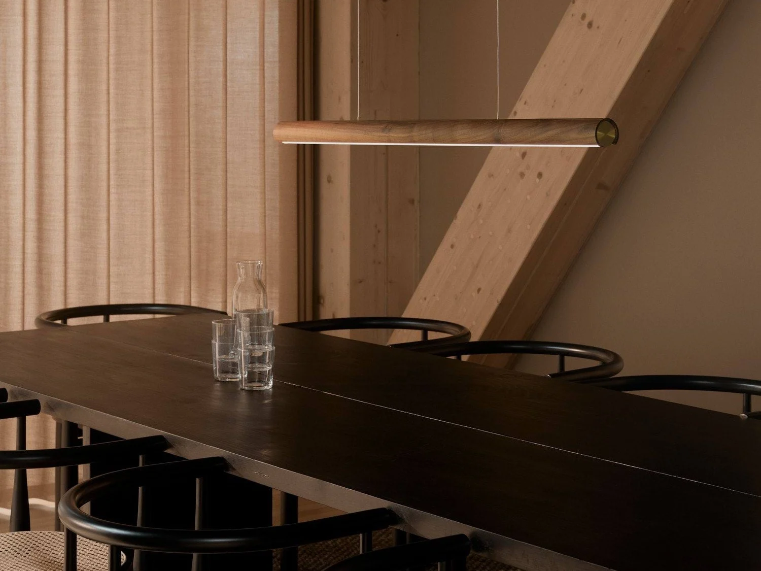 Une salle à manger moderne avec une longue table en bois foncé, des chaises noires et une élégante suspension en bois. Une carafe en verre et trois verres sont sur la table.