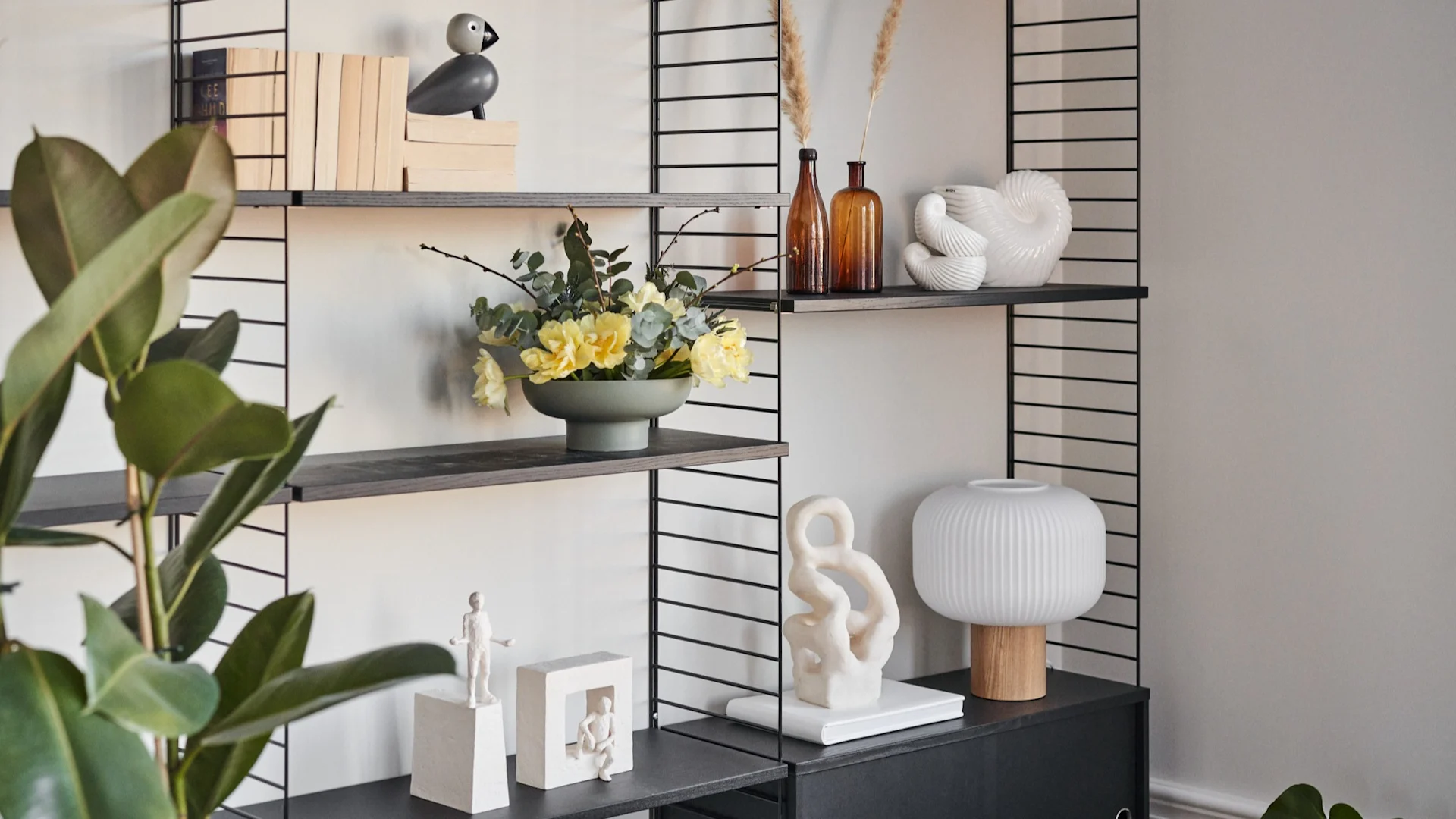 Étagère noire: livres, oiseau, fleurs jaunes, vases ambrés, sculptures blanches et lampe; plante verte en avant.