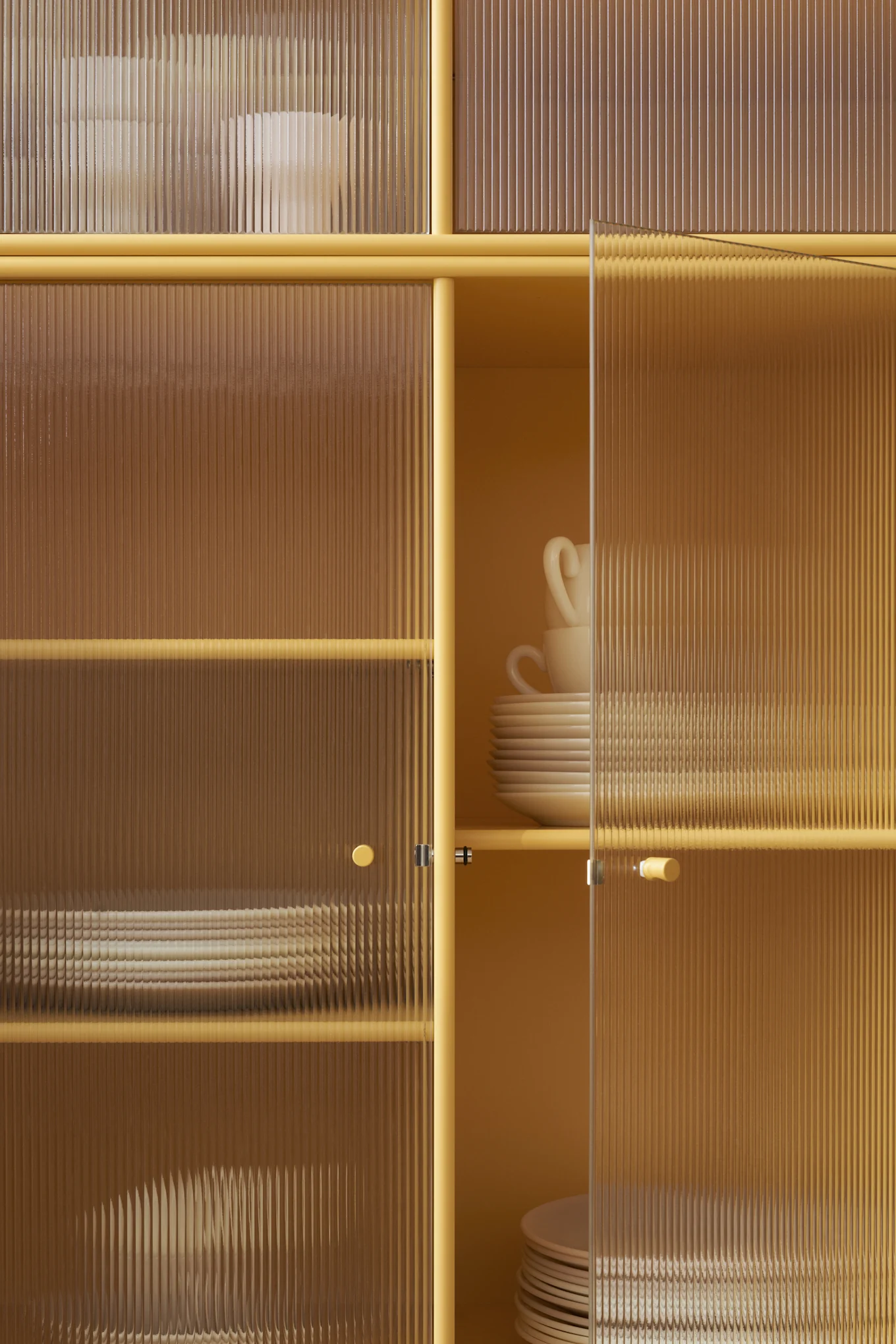 Meuble jaune moutarde avec portes en verre cannelé, révélant des assiettes et tasses blanches empilées à l'intérieur. Une porte est entrouverte.